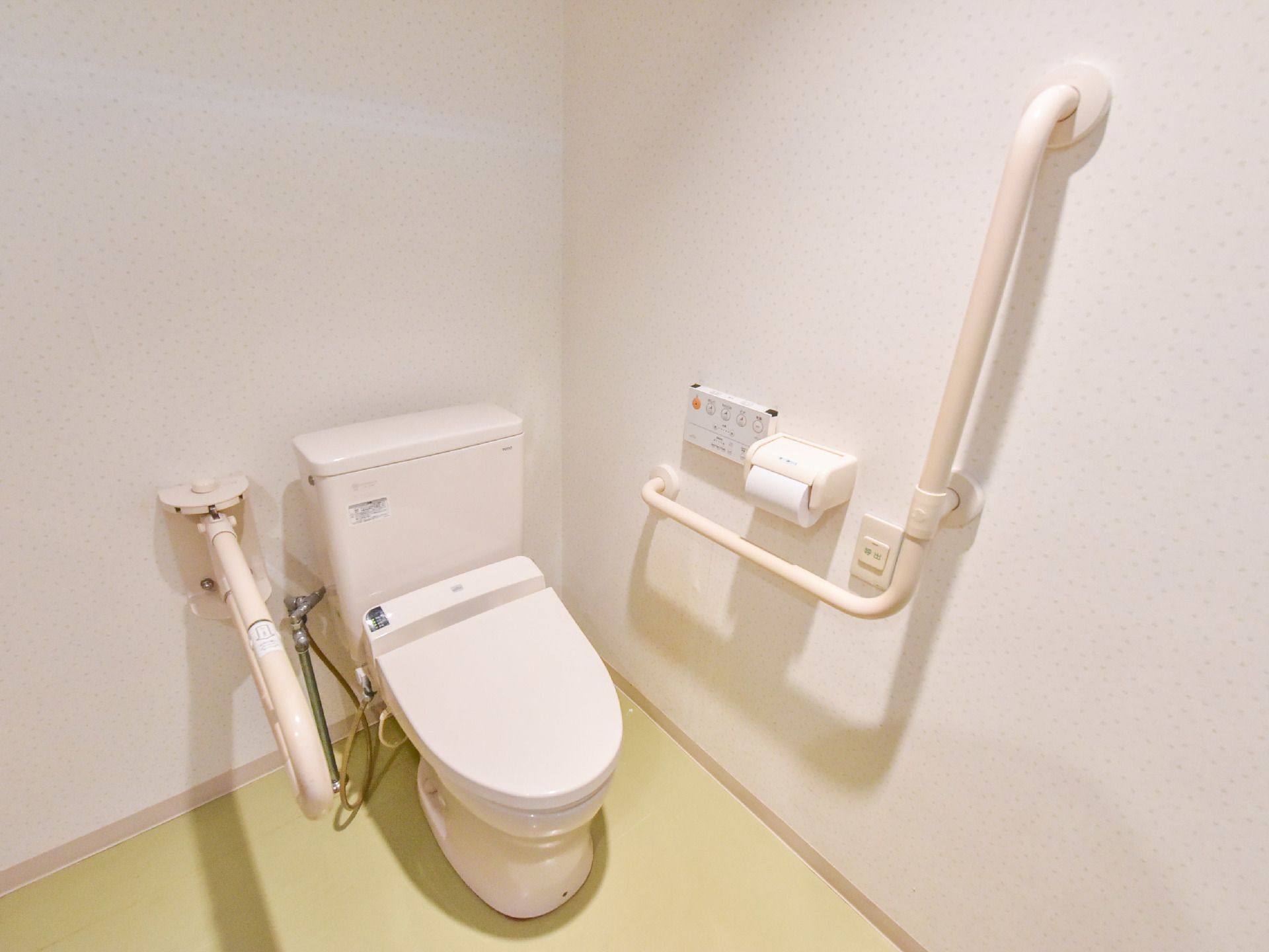 薄い黄緑色の床、白い壁のトイレには洋式便器がある。便器に座って右側と左壁側に大きさの違う手すりが付いている。