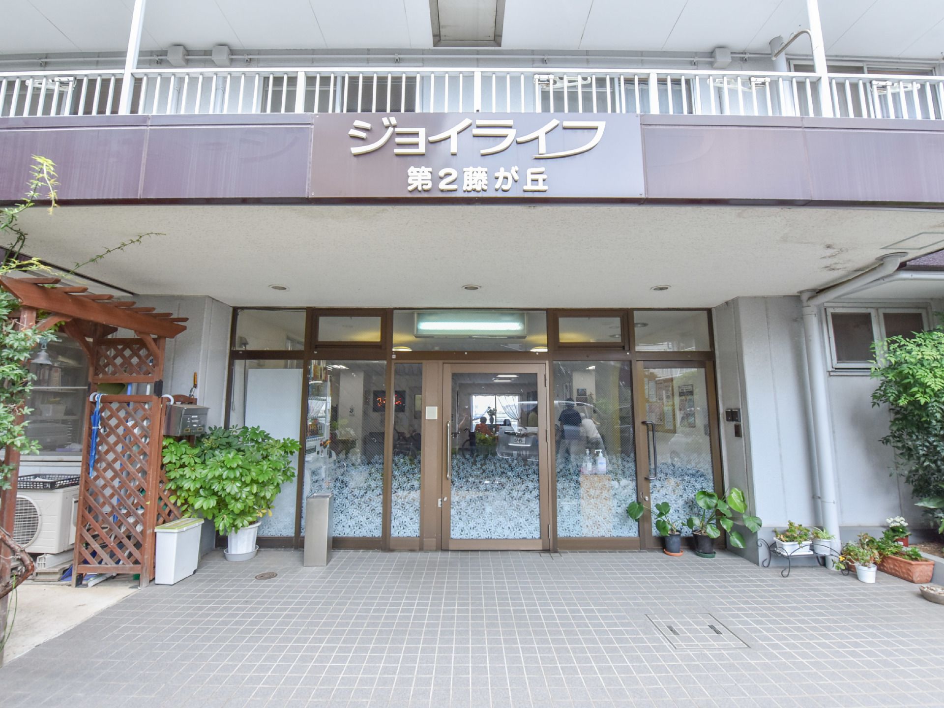 入口ドアはガラスになっていて中の様子が見える。2階のベランダが屋根になっている。玄関前には鉢に植わった植物がある。