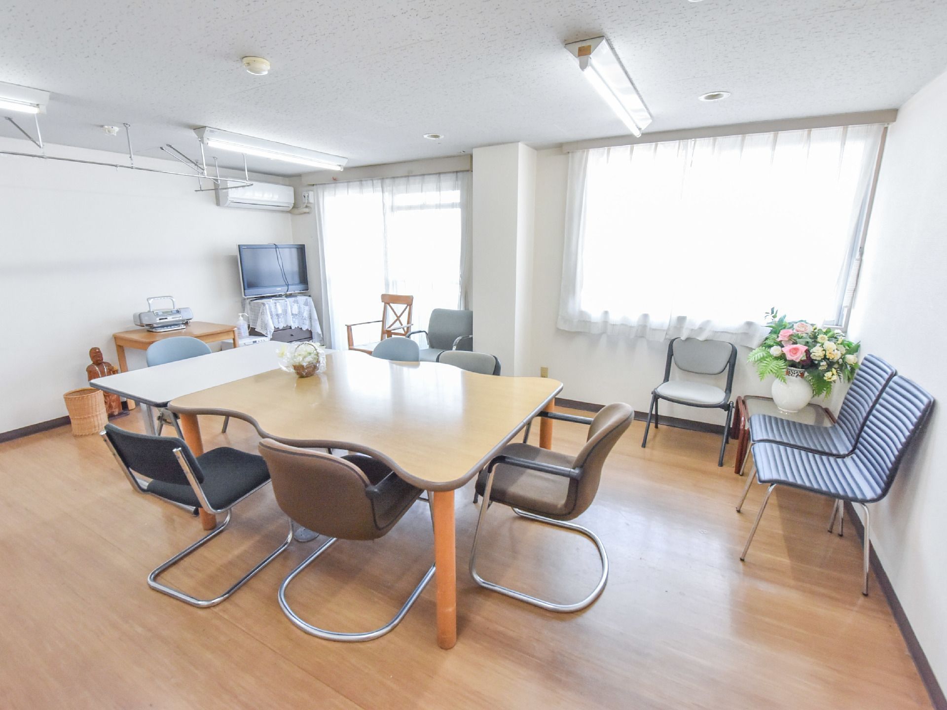 机と椅子が置かれたレクリエーションなどができる部屋で、窓から光が入る明るい空間で、お花が飾ってある。