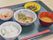 黒いお盆の上に赤いお箸がある。ご飯、お味噌汁、柔らかく煮たお魚の他、野菜のおかず2品の食事が用意されている。