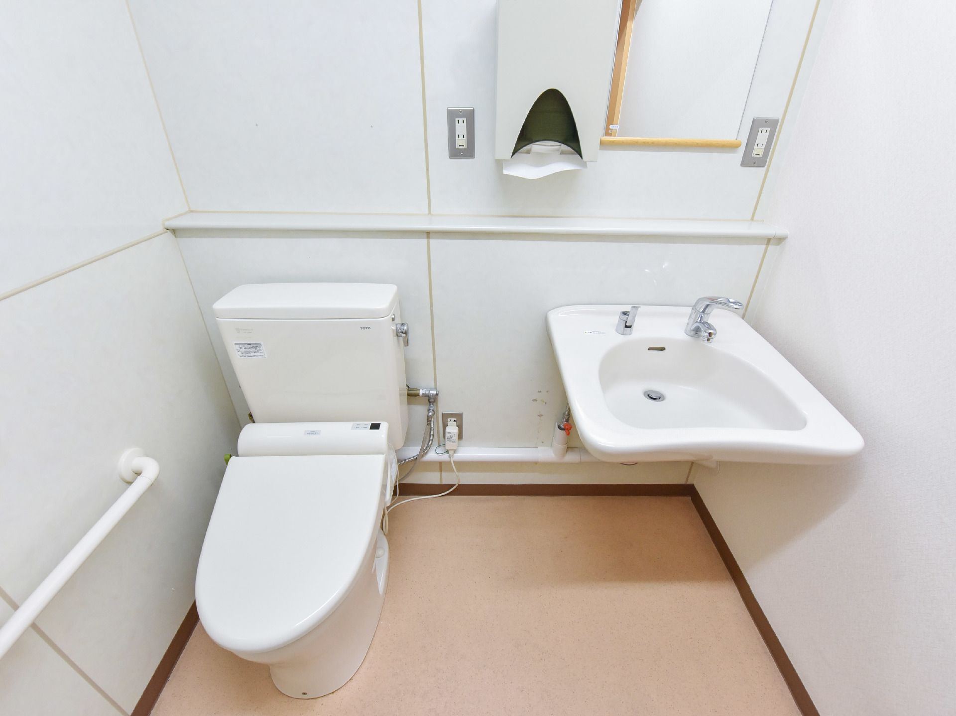 トイレの壁側には転倒防止の手すりが取り付けられている。すぐ横には鏡付きの洗面台も横に設置されている。