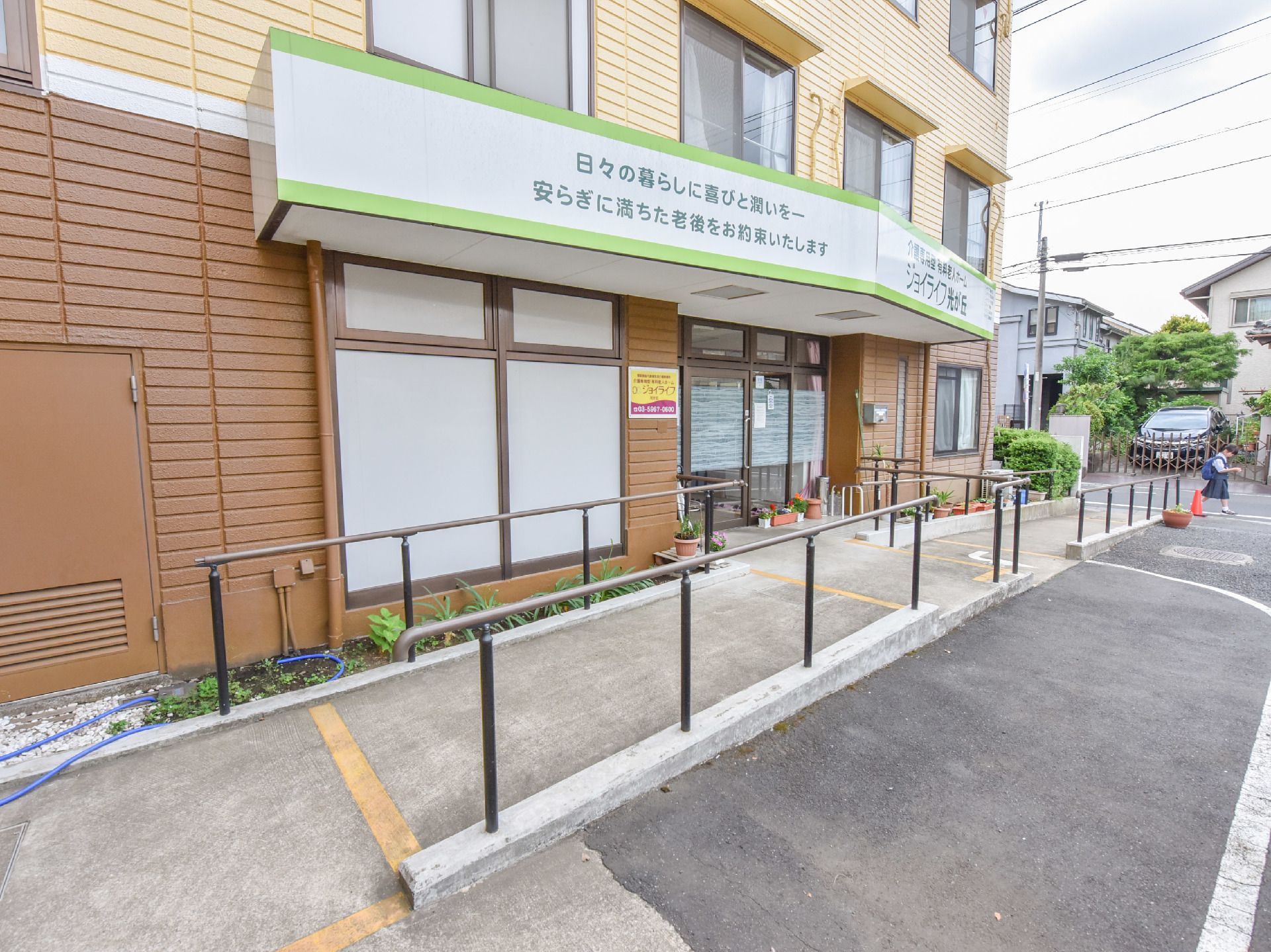 施設前玄関には施設名が大きく書かれた屋根がある。手すり付きのスロープがある。屋根の上には部屋の窓が見える。