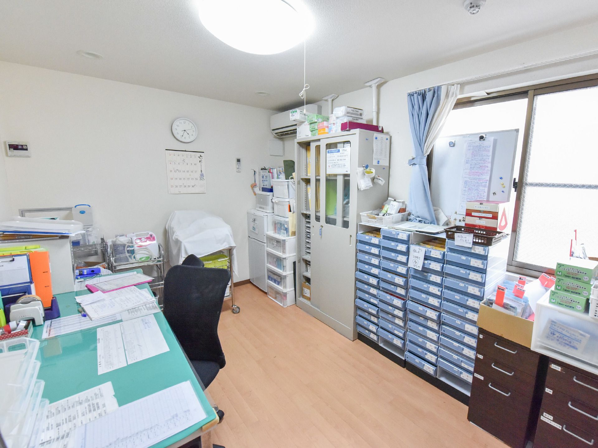 事務机と椅子が並んだ後ろ側には書類収納棚やロッカーがずらりと並べられている。壁には丸い時計、カレンダーもある。