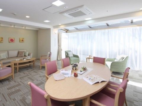 丸テーブルの周囲には椅子が置かれており、入居者様同士などでお茶など楽しめる。奥には広い窓があり、柔らかい日の光が差し込んでいる。
