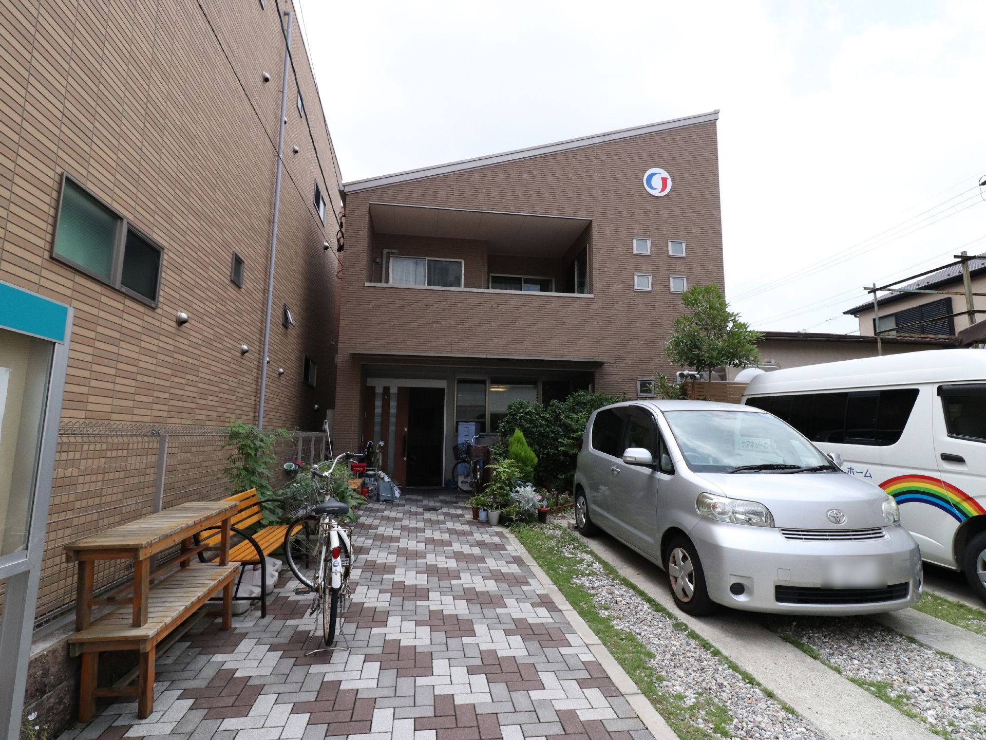 ブラウンの外壁で2階建ての建物になっている。玄関まではレンガが敷かれ、木々が植えられているので自然を感じることができる。