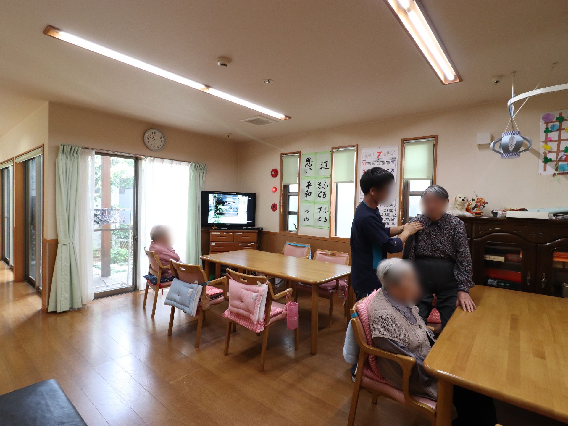大きな窓から光が入り、白い天井と壁で明るい部屋になっている。壁には、時計やカレンダー、入居者の書道の作品が飾ってある。