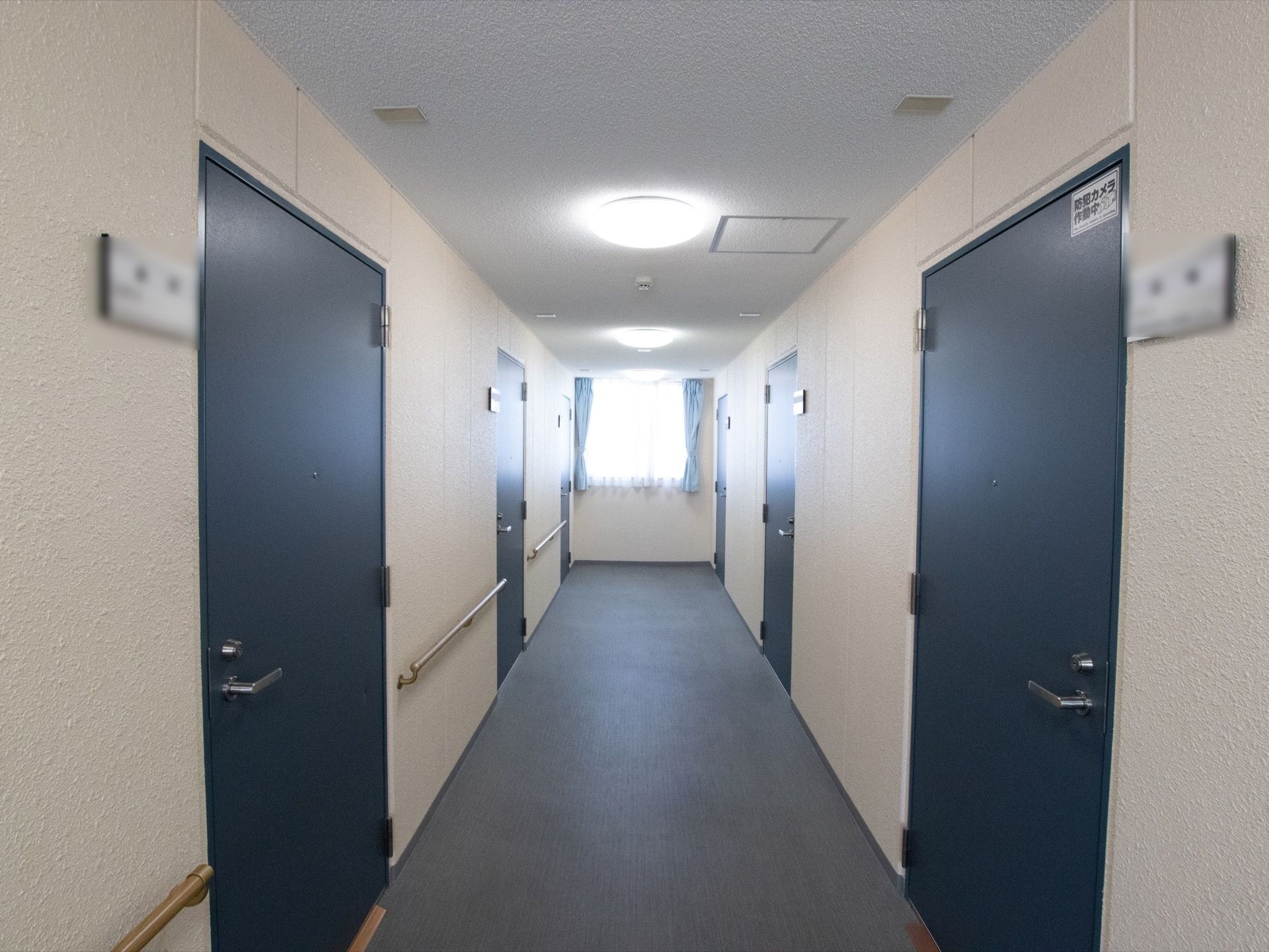 各居室の入口は濃い青色の片開きのドアになっている。ドアの横に表札が貼られている。廊下の奥に窓がある。