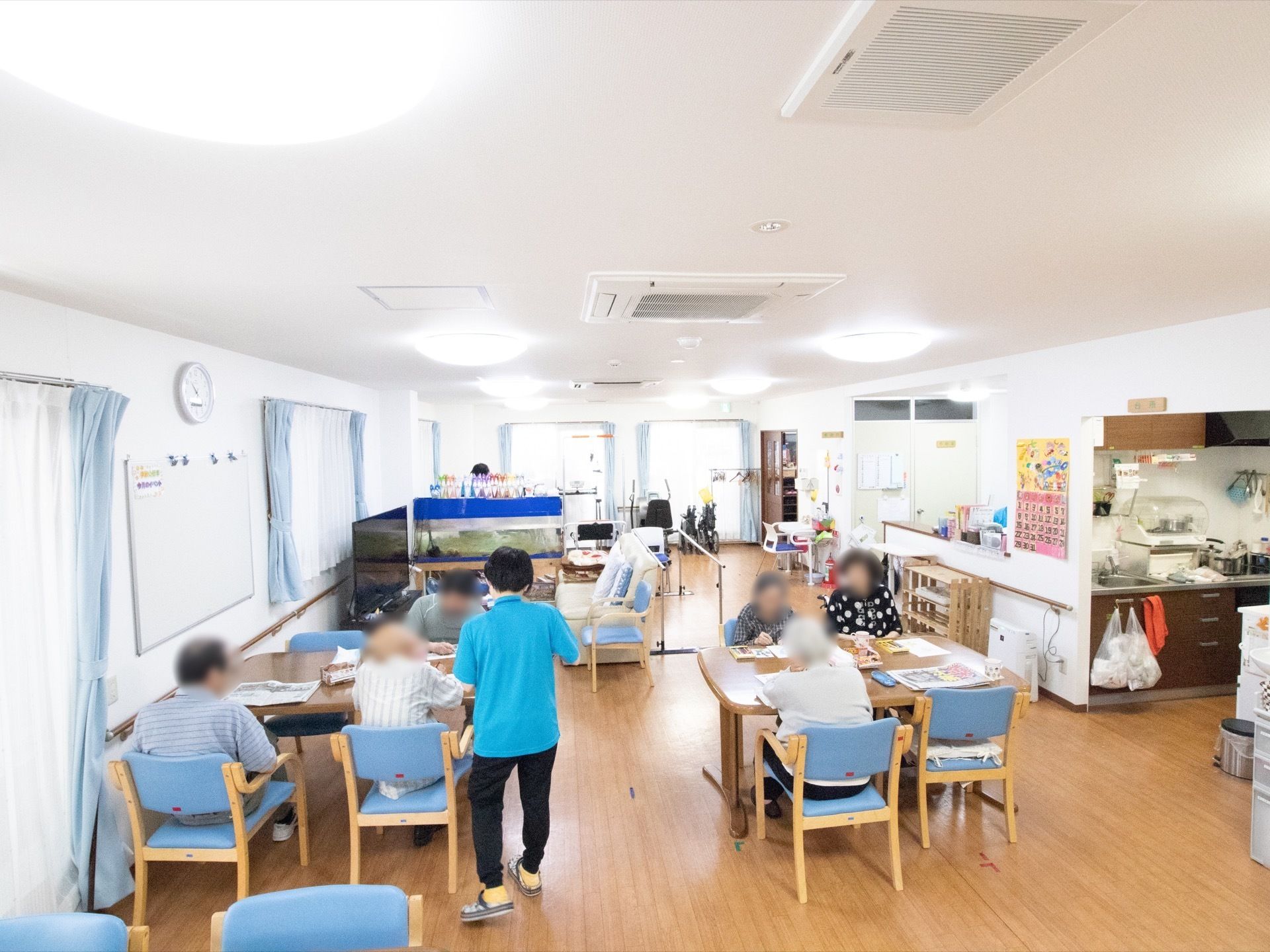 塗り絵をされている入居者様や新聞を読まれている入居者様がいる。ポロシャツを着た職員が入居者様とお話をしている。