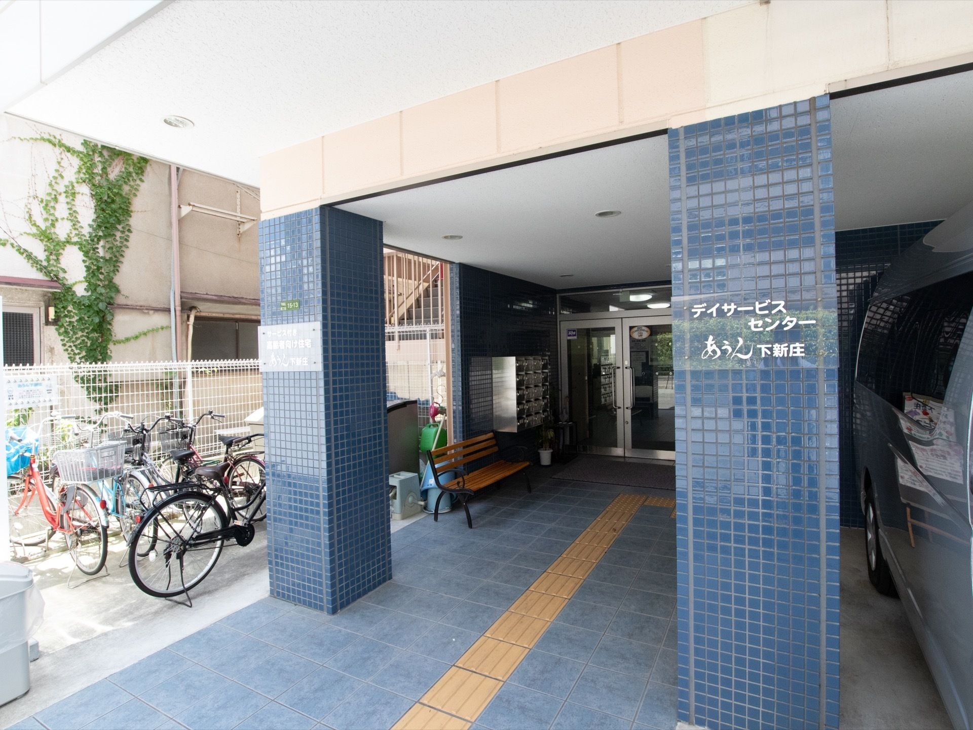 青いタイル状の柱に施設名と住所が書かれている。建物の左側には駐輪場があり、たくさんの自転車が停められている。
