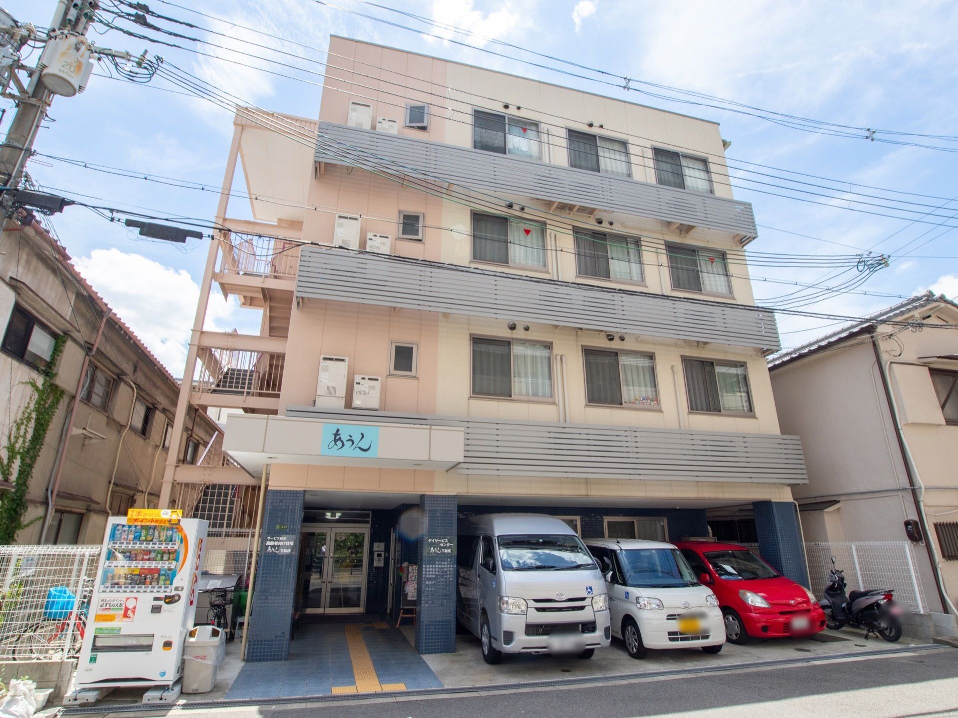 ４階建ての建物で、施設前に車が３台並んでいる。入口近くには自販機が設置されており、隣にはゴミ箱が用意されている。
