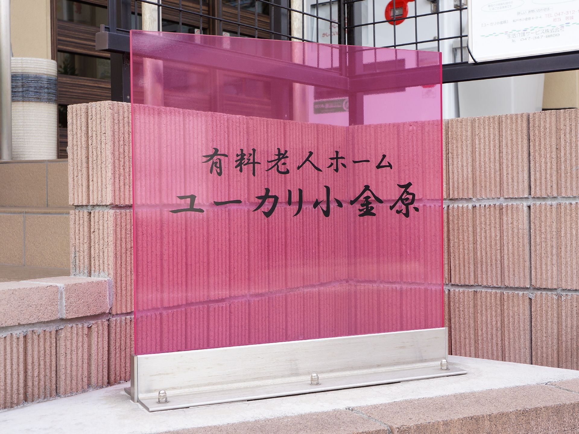 ピンク色のアクリル板に施設名が黒色で描かれている。茶色の塀の前にたっており後方に階段と入り口が見える。