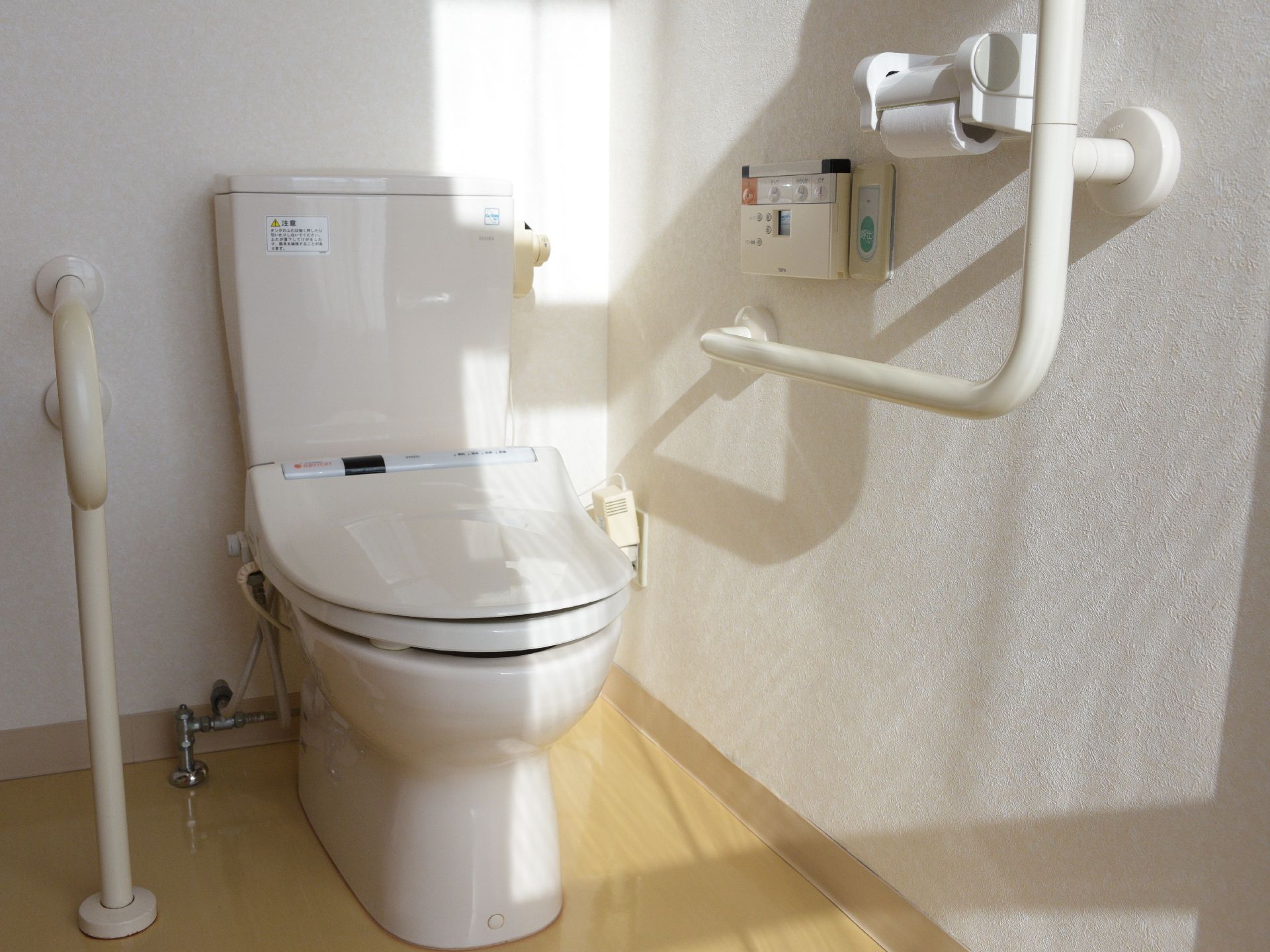 白い壁に薄いベージュの床のトイレである。手すりが体の両側に設置されているので安全に利用できる。温水洗浄便座になっている。