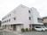 施設は３階建てで、薄いベージュに近い色である。外壁の上部には施設名が書かれている。施設の周りには植え込みがある。