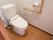 清潔感のある白い洋式トイレである。壁には温水洗浄機能の操作ボタンが取りつけられ、手すりも設置されている。