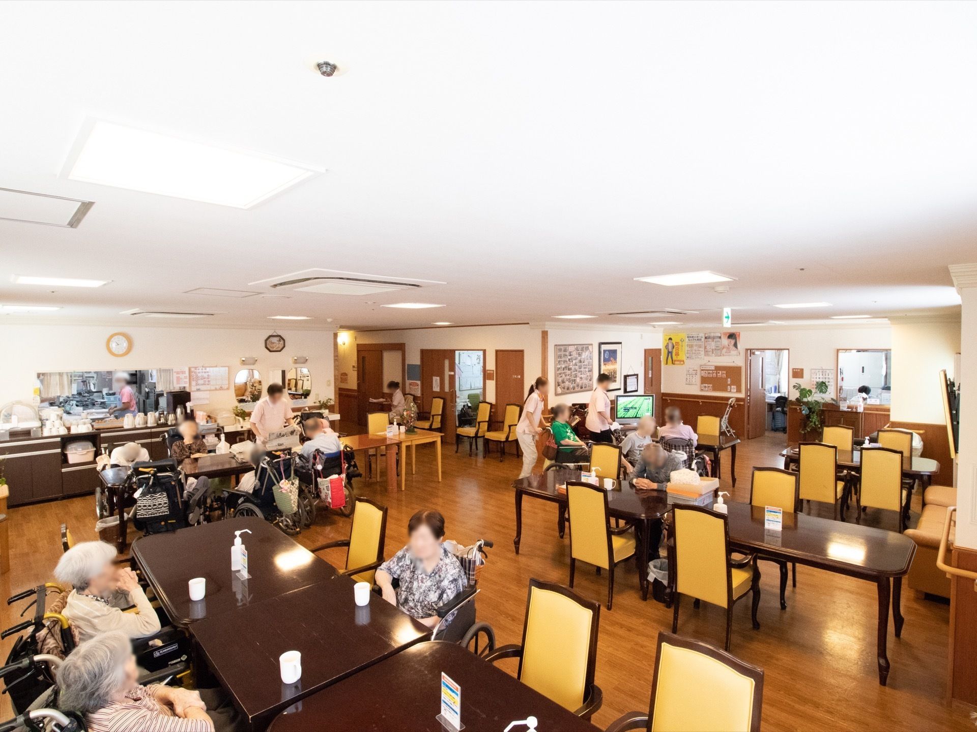 ダイニングテーブルや椅子が置いてある部屋は、たくさんの入居者様でにぎわっている。入居者様は椅子や車椅子に腰を掛けている。