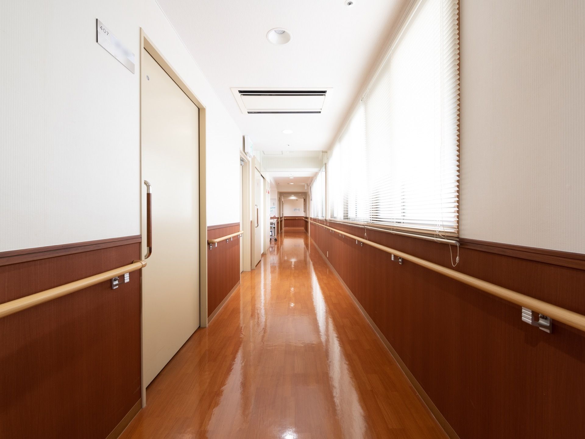 ぴかぴかに磨き上げられた廊下は、左側に居室のドアが並んでいる。右側は窓が続き、ブラインドが下げられている。