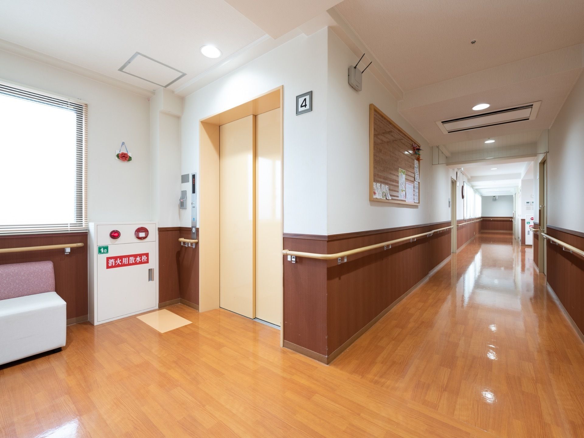 右奥に廊下が伸び、壁に大きな額縁が飾られている。左手前にエレベーターがあり、壁際に椅子が置かれている。