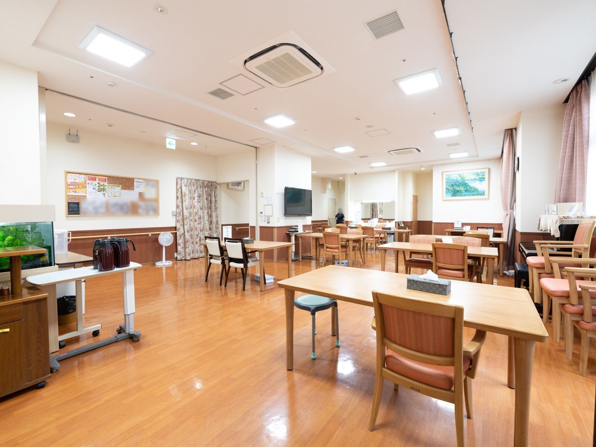 ダイニングの奥の柱に大型液晶テレビが取りつけられている。壁には掲示板が貼られ、施設からのお知らせなどが掲示されている。