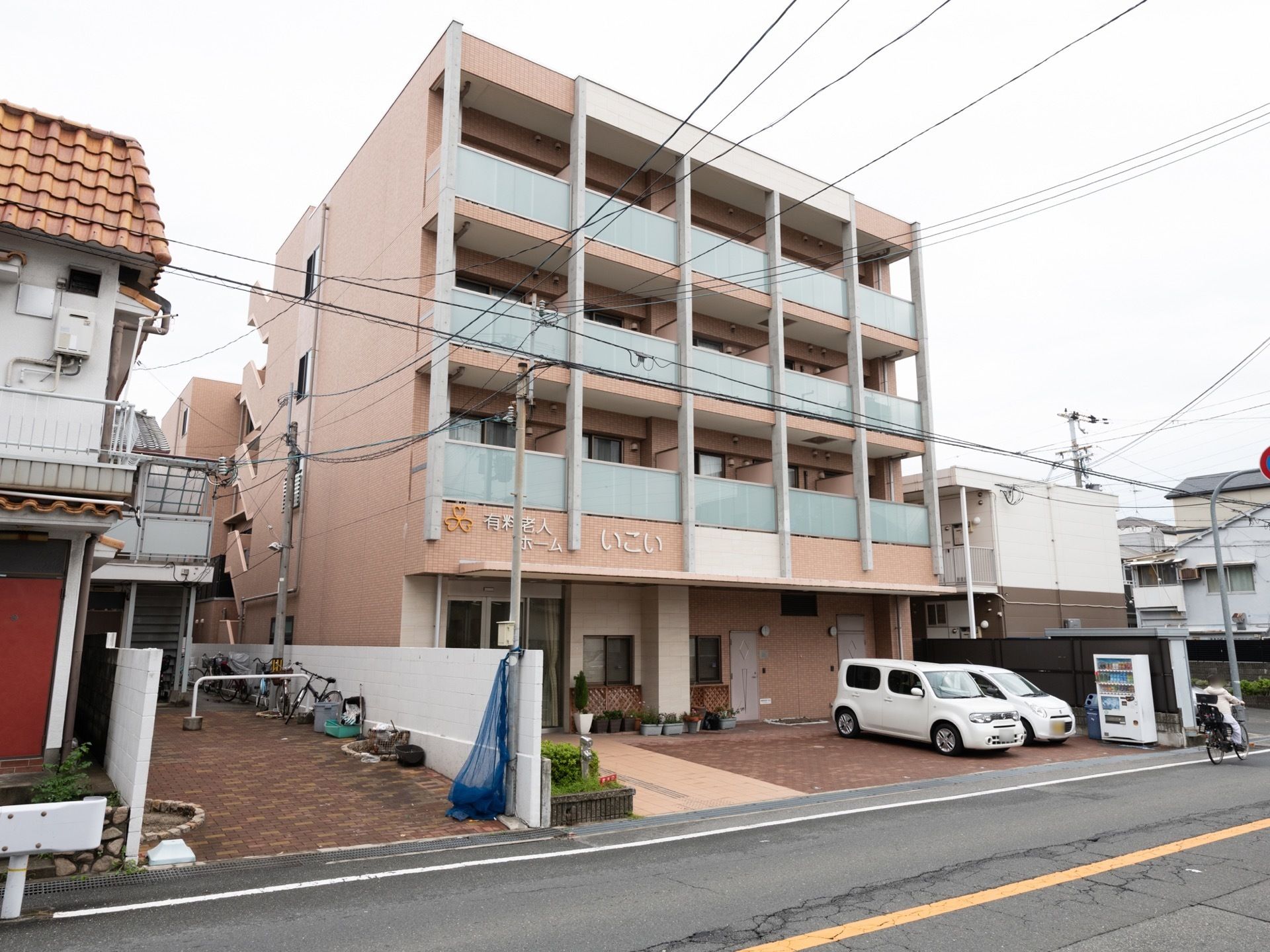 施設の壁は淡いオレンジ色で、ベランダが半透明の板になっている。通りに面しており、施設前には車が２台停まっている。