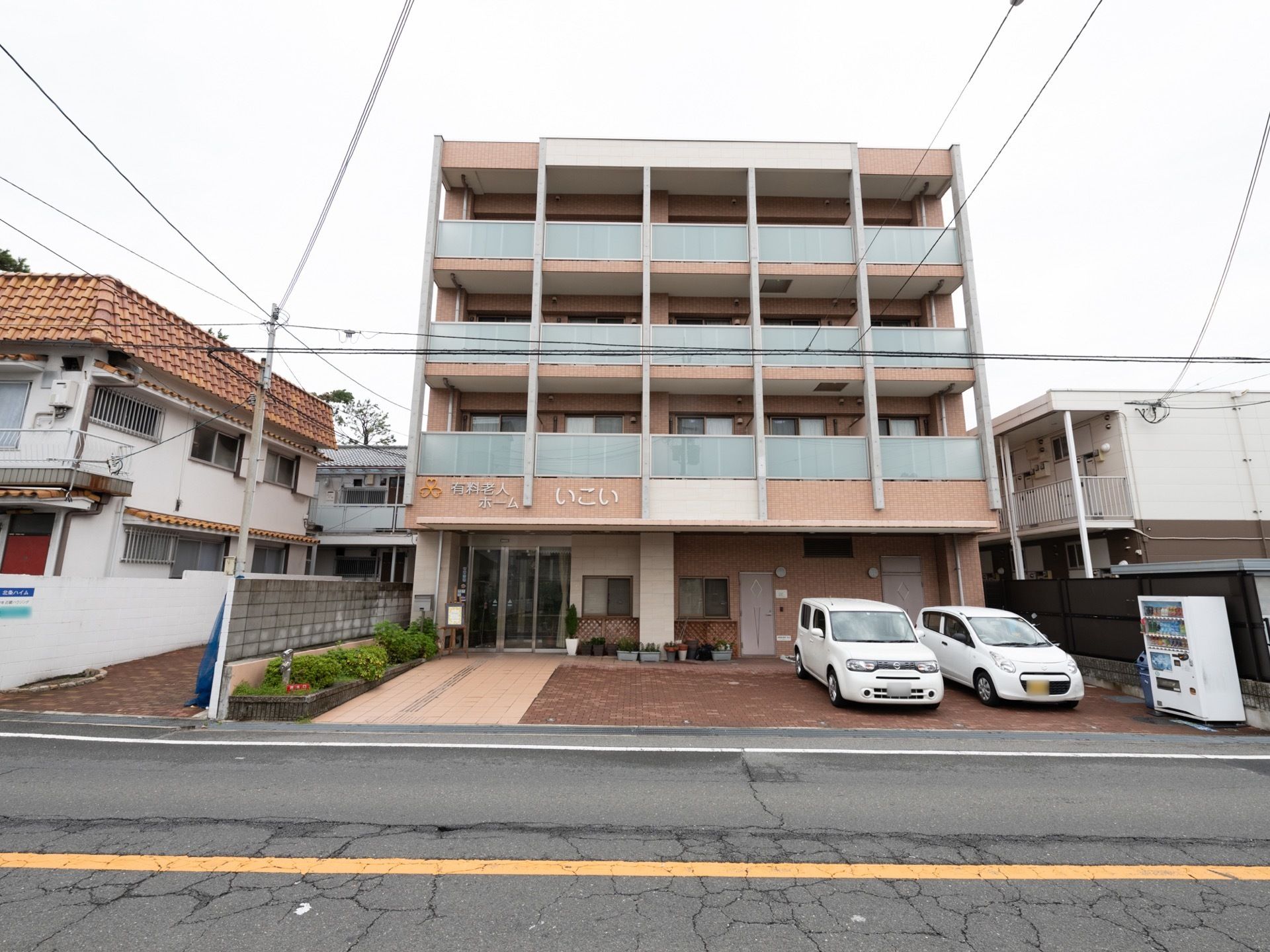 建物の前には道路が通っていて、敷地の一角には自動販売機が設置されている。道路から玄関へ向けて植え込みが作られている。
