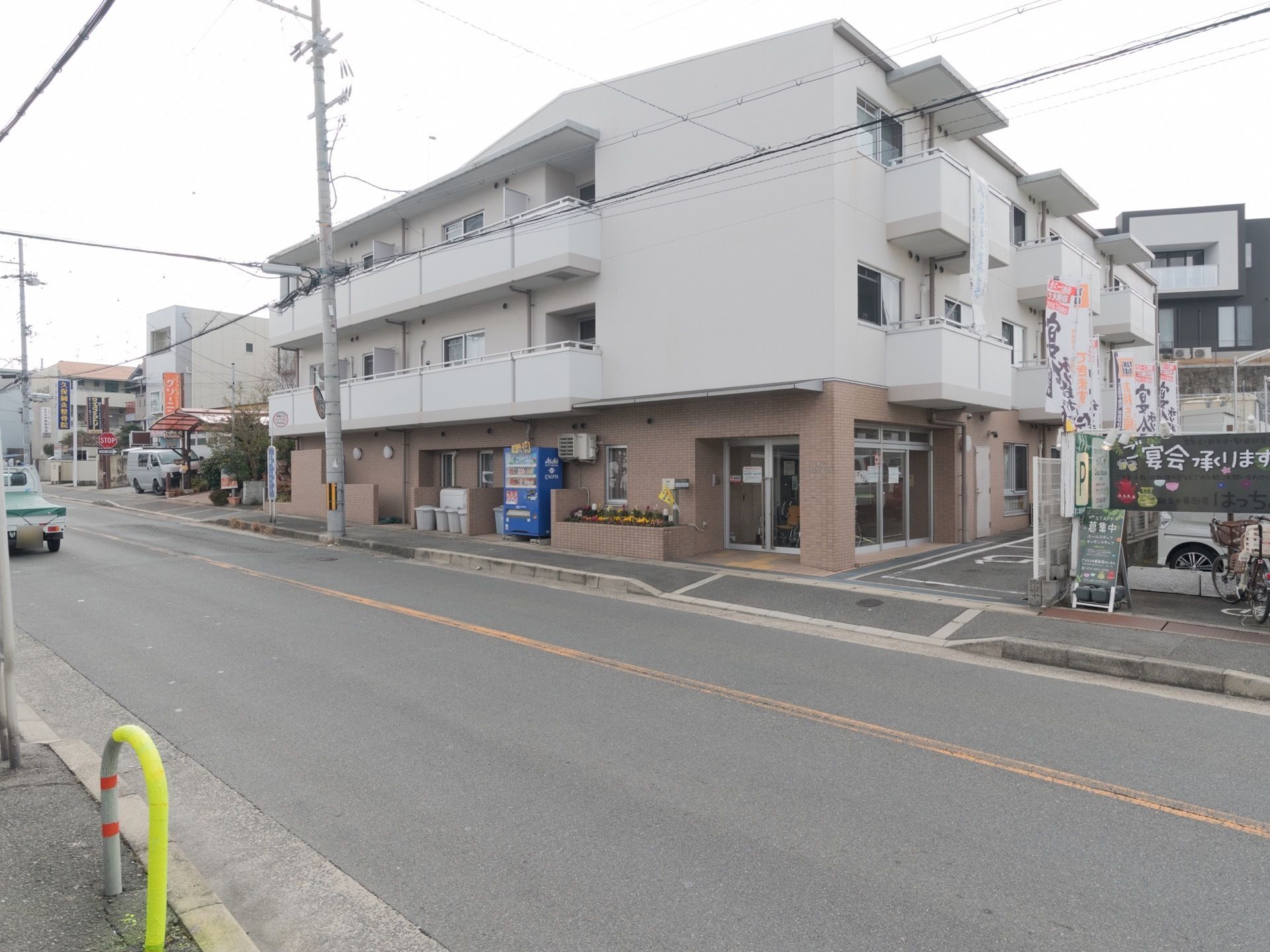 幹線道路に面して施設建物が建っている。3階建てで道路に面した部屋には隣室とつながったベランダがある。