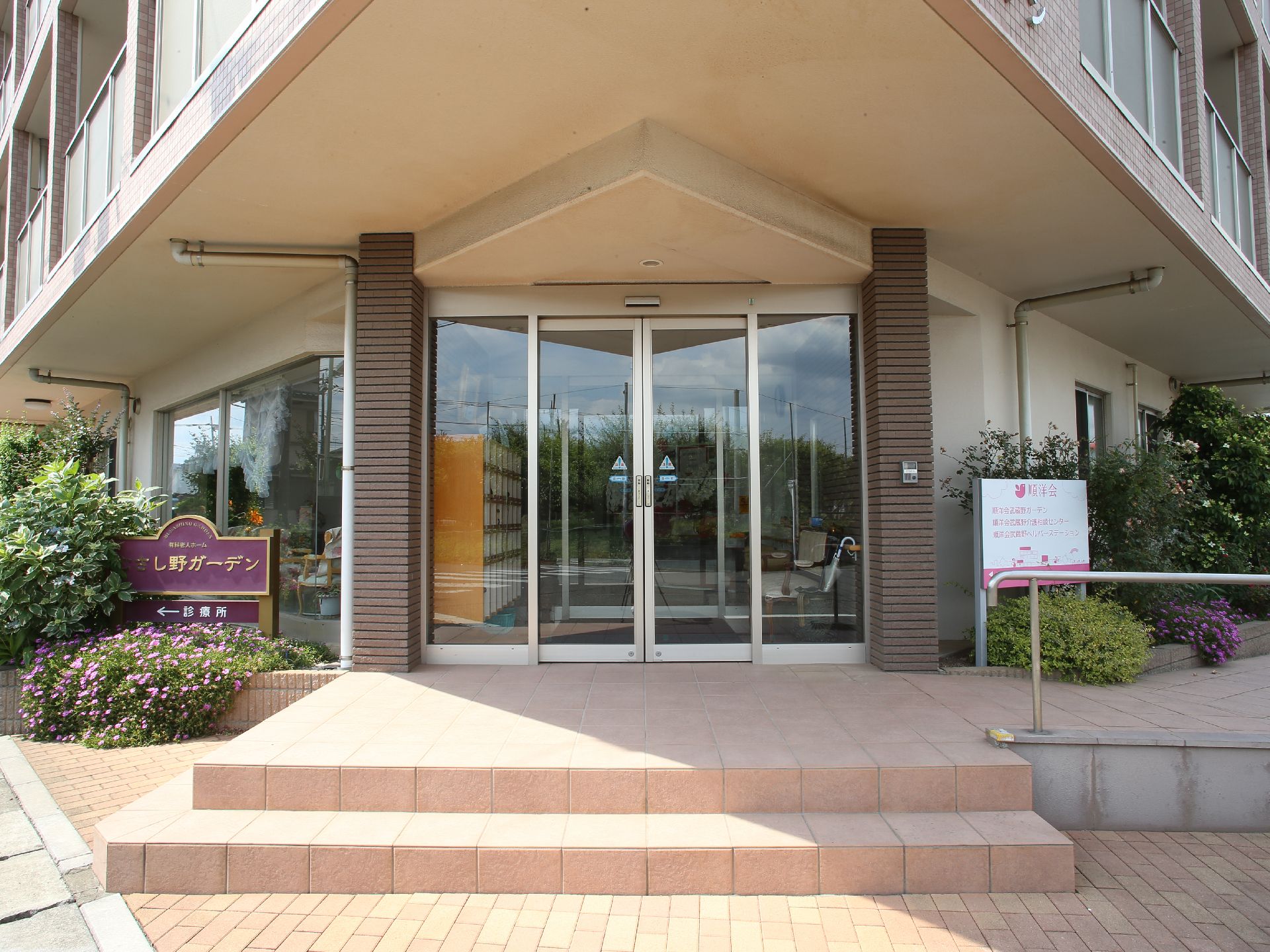 玄関の前に階段があり、手すりが備わったスロープも設けられている。大小さまざまな植栽が植えられている。