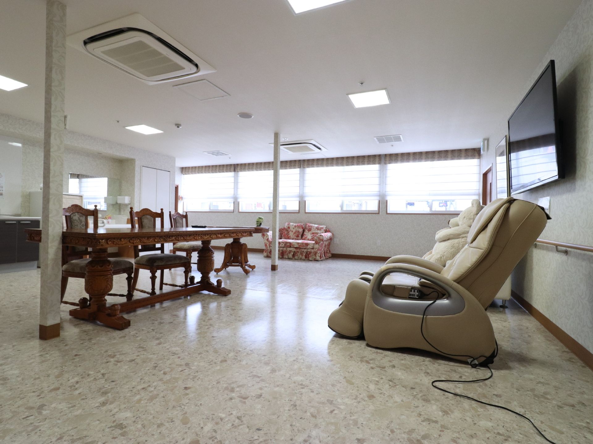明るい広いお部屋でイスやテーブル、ソファがある。テレビの下にマッサージチェアが設置されていて、ゆったり座ってリラックスができる。