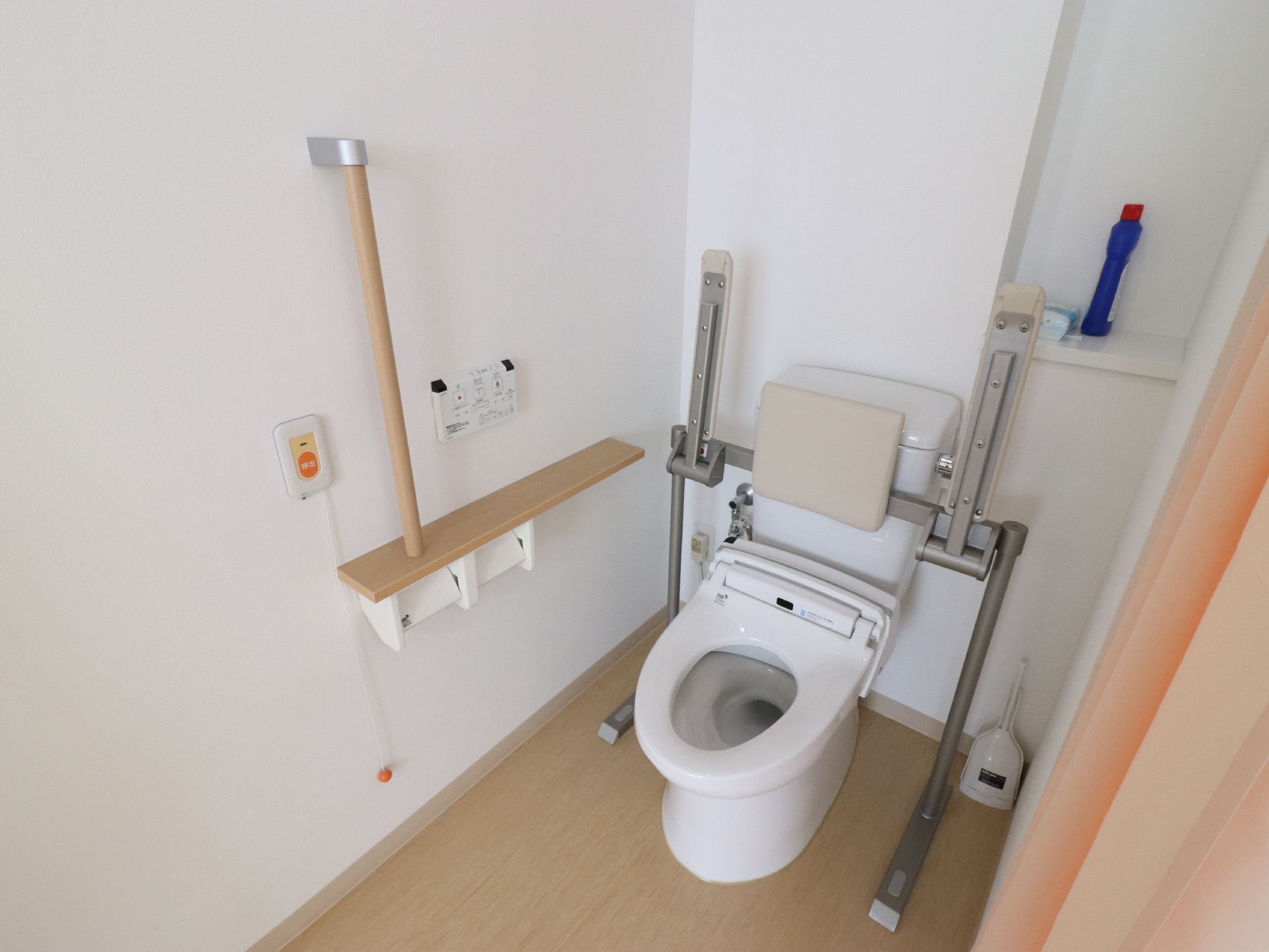 各部屋のトイレである。木目調で、壁には手すり、背もたれ、肘置き、緊急用のコールボタン、洗浄機能付き便座が設置されている。