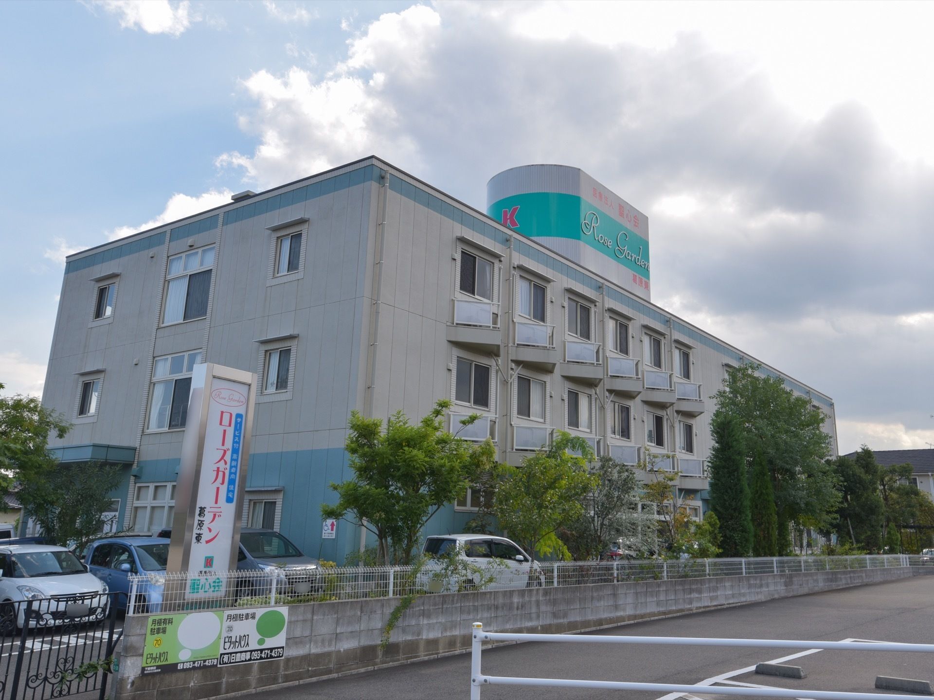 施設は3階建ての大きな建物で、建物の上にエメラルドグリーンの表札と駐車場に縦長の表札が設置されている。