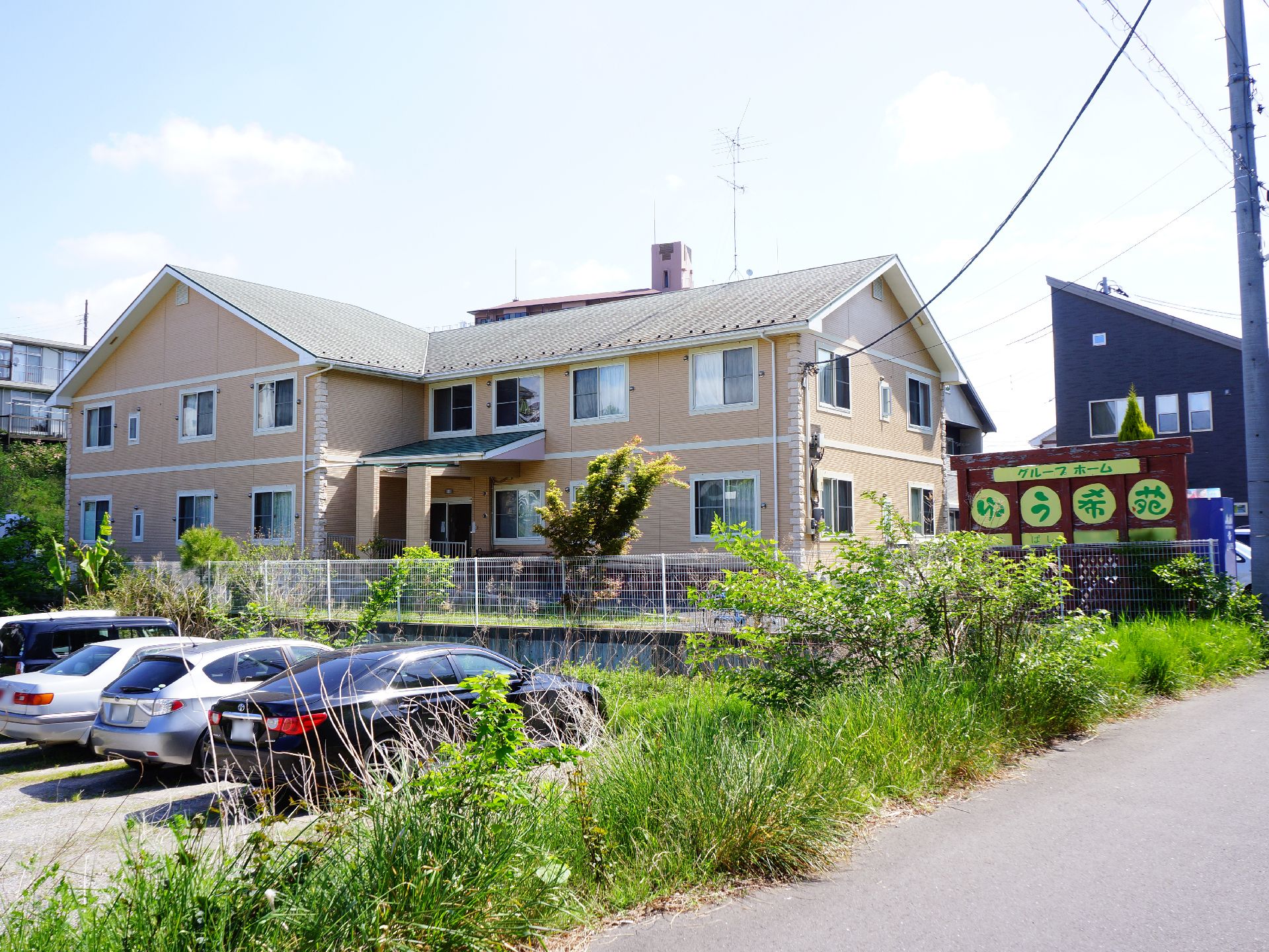 閑静な住宅街に位置するクリーム色の外壁の鉄骨造2階建の外観。敷地内入口の所に道路から見やすい様に看板が掲げられている。