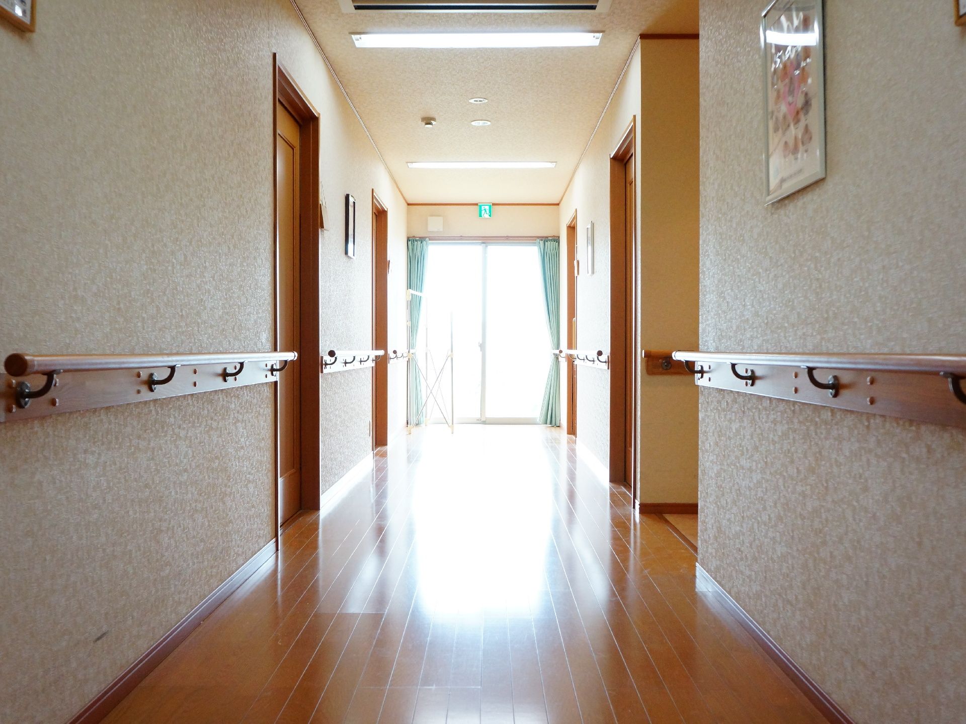 施設の建物内の廊下の様子。手すりが伸びる中に居室などの扉が並ぶ。採光に使われている一番奥のガラス戸は非常口も兼ねている。