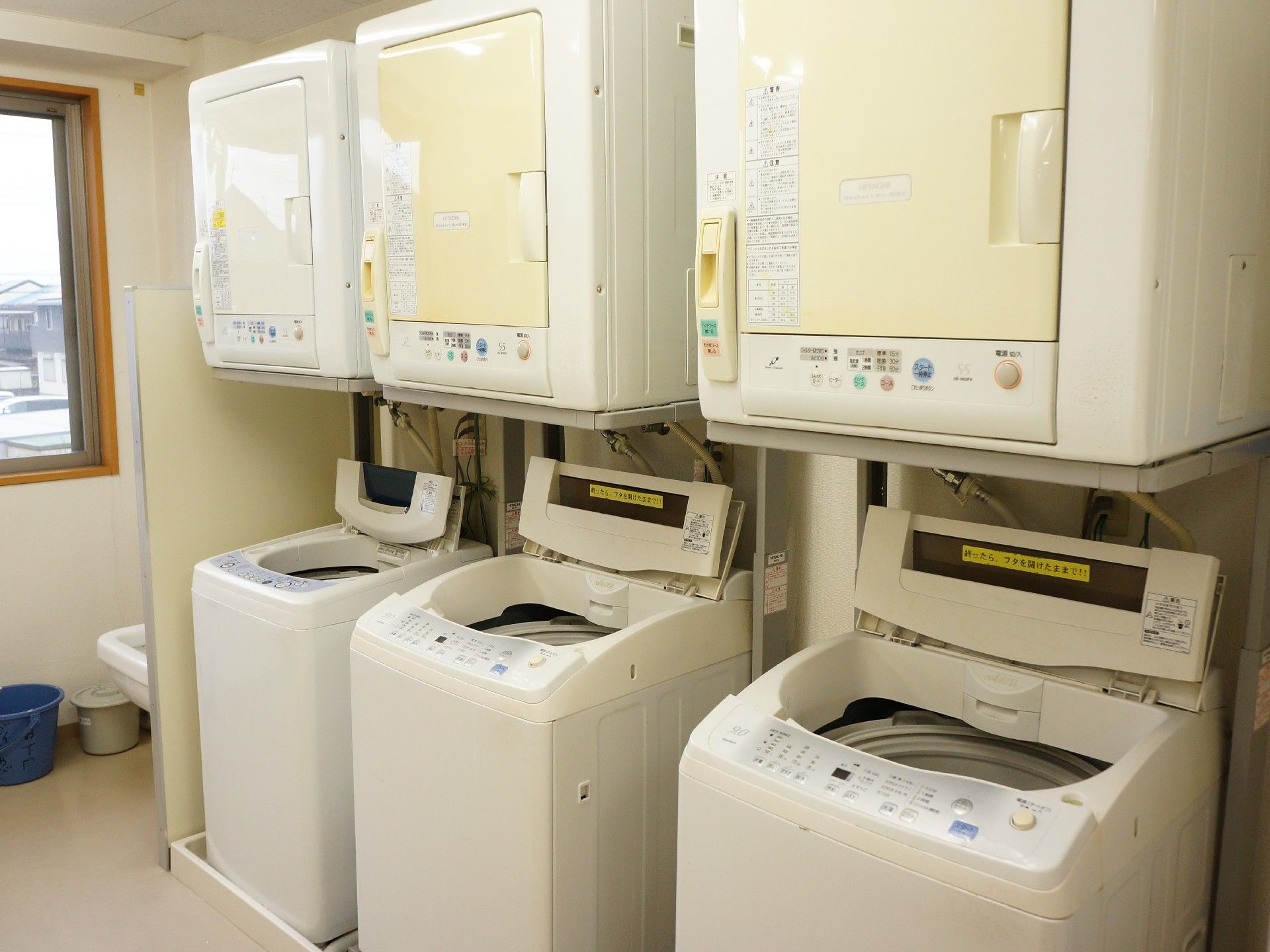 白を基調とした清潔感のあるランドリー室で、洗濯機と乾燥機が３台ずつ設置されている。小窓の下にバケツが置かれている。