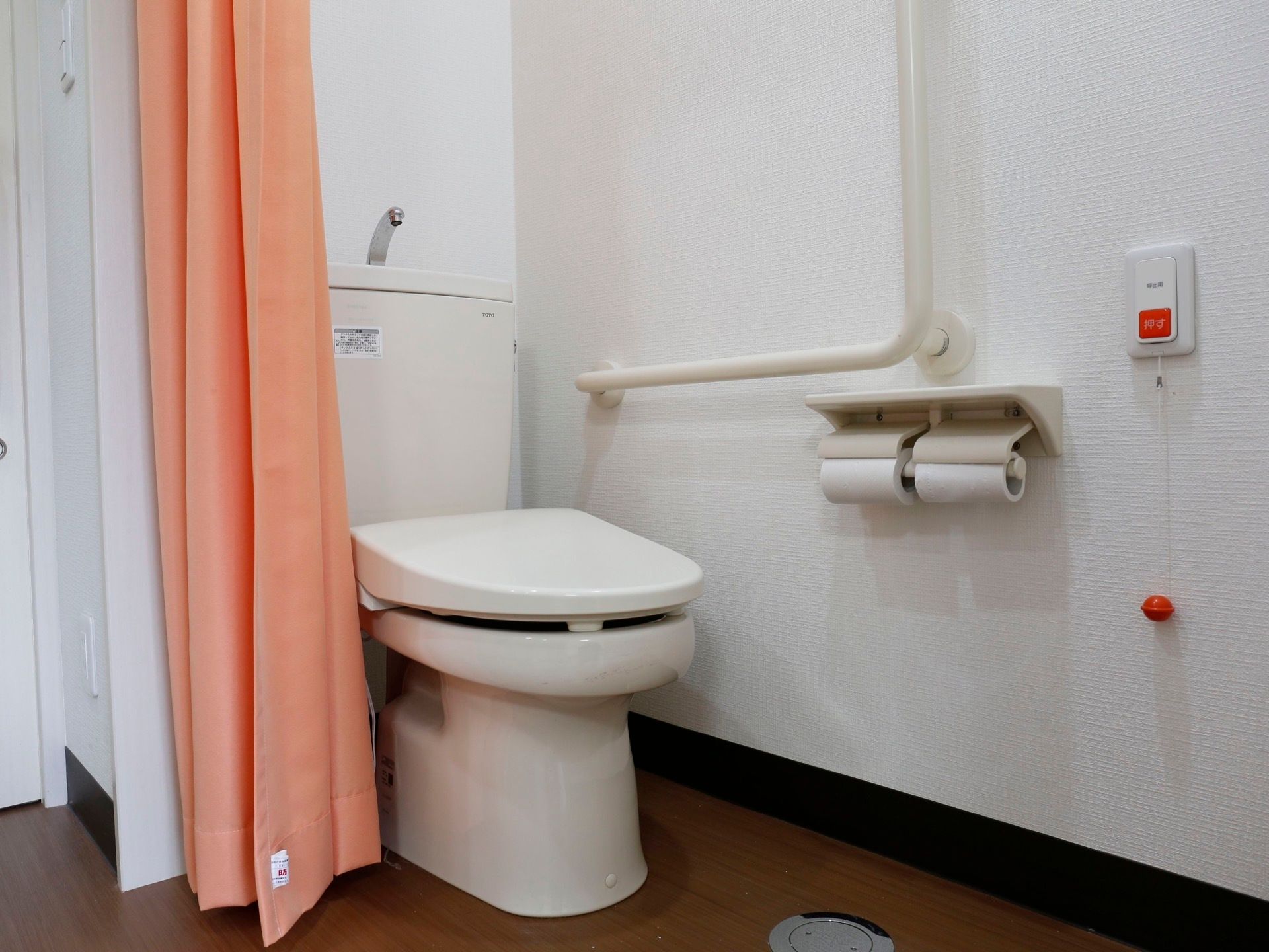 サーモンピンクのカーテンの向こう側にタンク式の洋式トイレが設置されている。壁に２連のペーパーホルダーと非常ボタンが設置されている。
