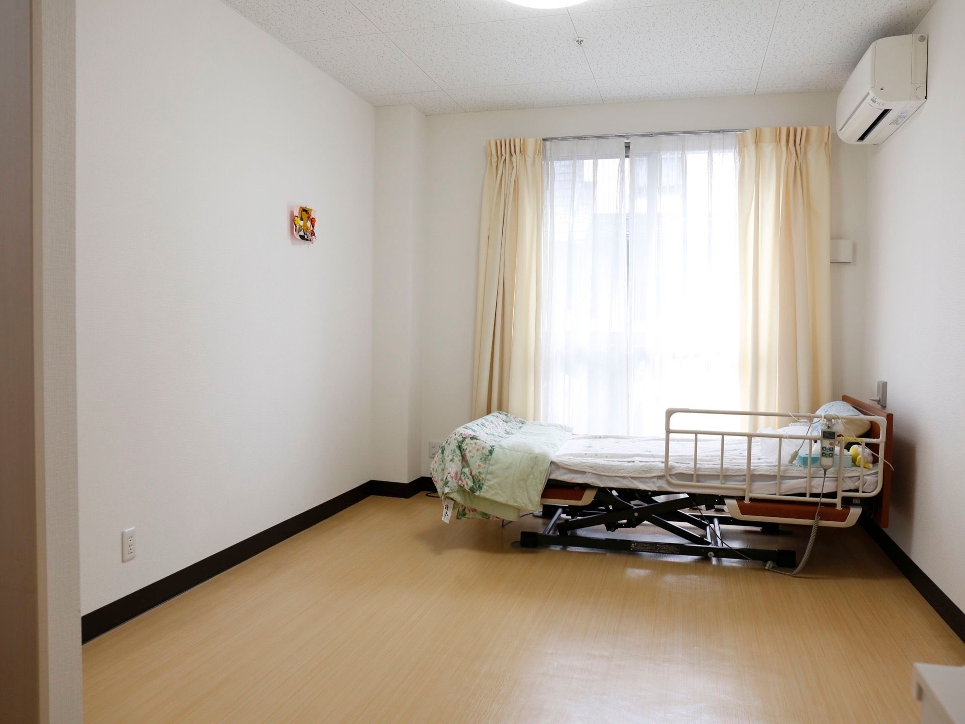 掃き出し窓にレースカーテンとクリーム色のカーテンが掛かっている。壁にはエアコンが設置され、転落防止柵付きのベッドがある。