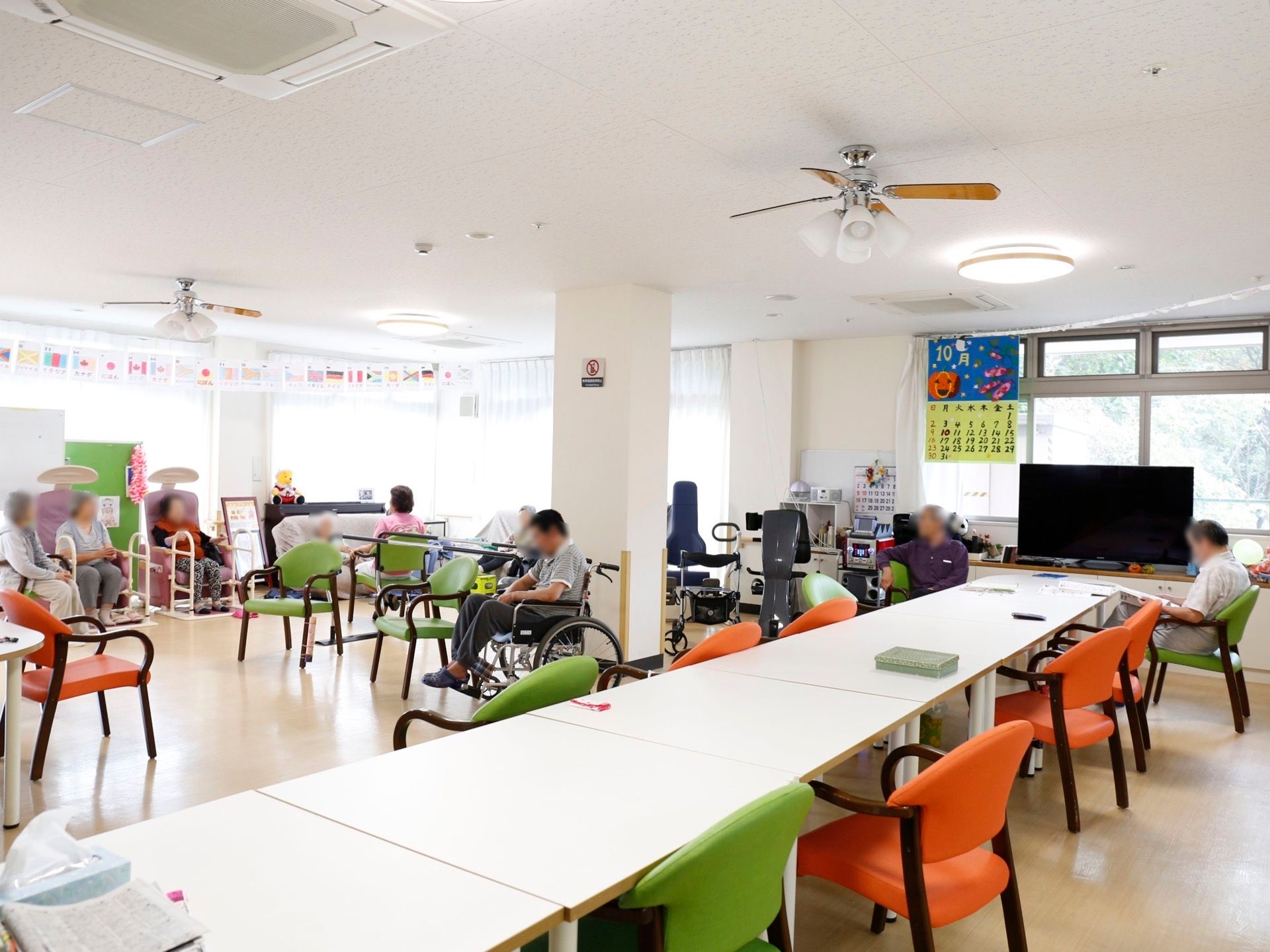 窓際に薄型テレビが設置され、大きなカレンダーが掛けられている。白いテーブルとオレンジ色と緑色の椅子が数脚置いてある。