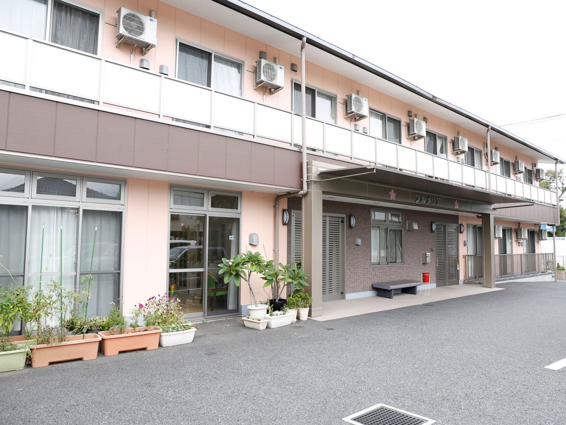 舗装された敷地に２階建ての淡いピンク色の建物が建っている。プラスチックのプランターや植木鉢に植物が植えられている。
