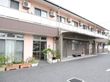 舗装された敷地に２階建ての淡いピンク色の建物が建っている。プラスチックのプランターや植木鉢に植物が植えられている。