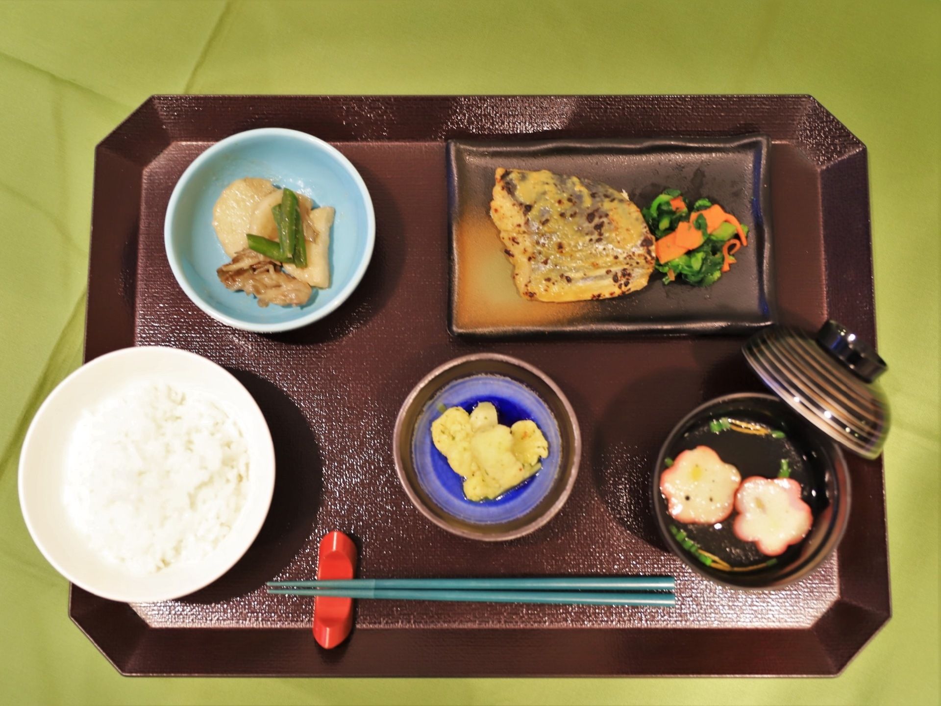 緑色のテーブルクロスの上に料理が並べられている。魚料理とお吸い物があり、健康的でバランスの取れたメニューとなっている。