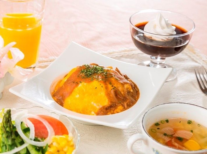 オムライスを中心とした洋食の食事である。テーブルにはテーブルクロスが掛けられており、ほかにはスープやサラダ、コーヒーゼリーが用意されている。