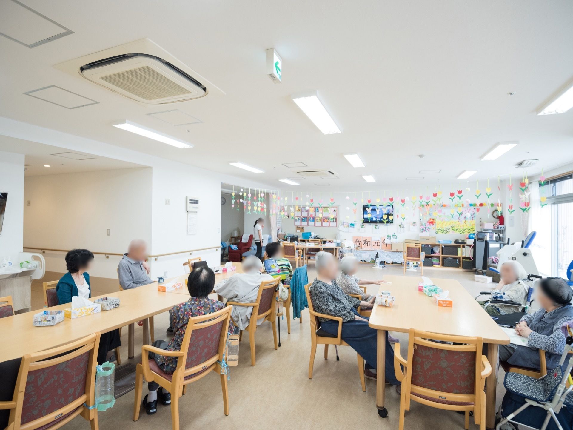 テーブルと椅子が並べられている広い談話室がある。入居者様達が大勢で集まっている。天井からは飾りつけが下がり、壁際には棚が置かれている。