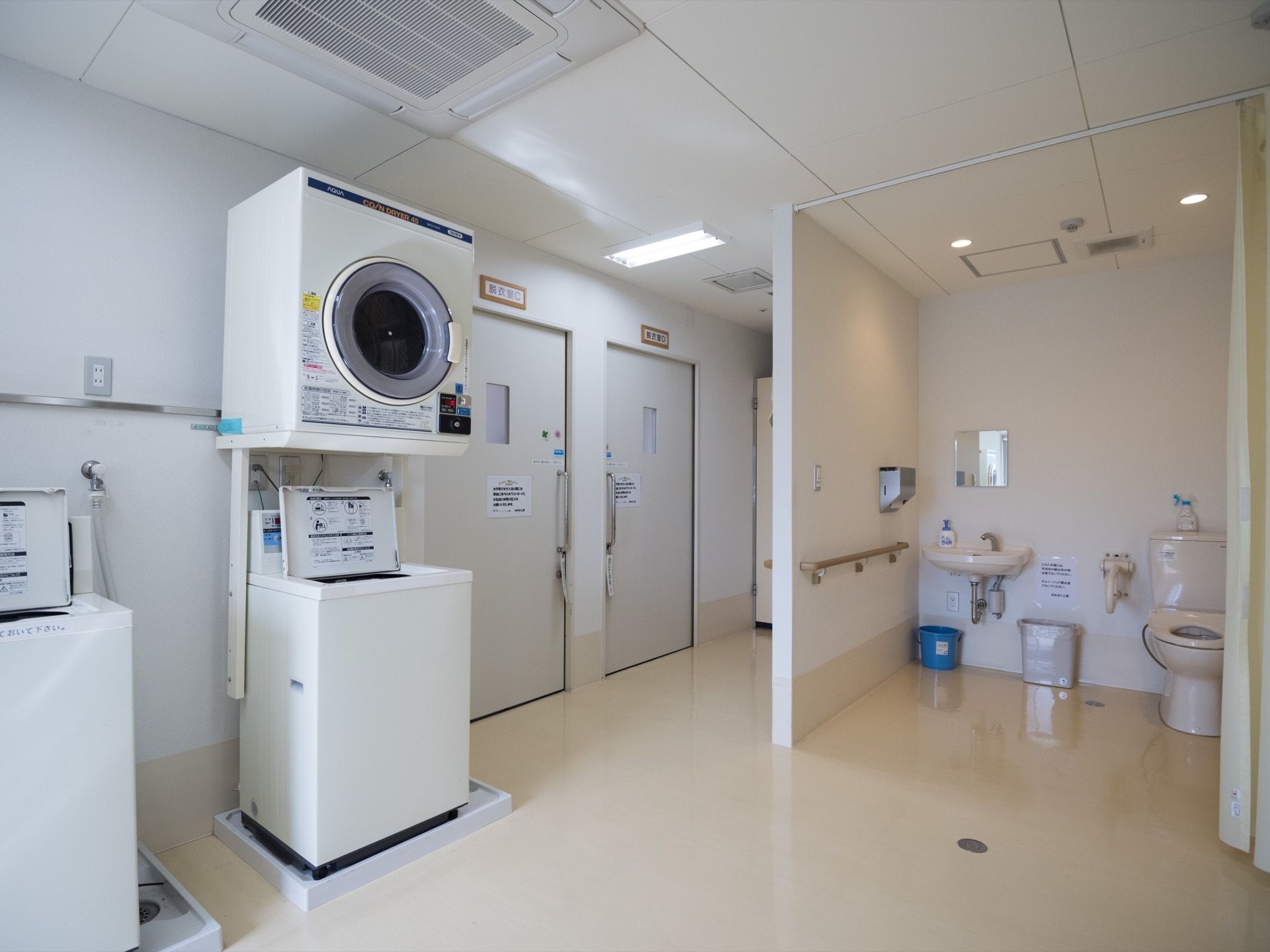 カーテンの掛かった広いトイレの前に洗濯機が置かれている。そのうちの１台の上には乾燥機がある。天井には大きな空調設備が設置されている。