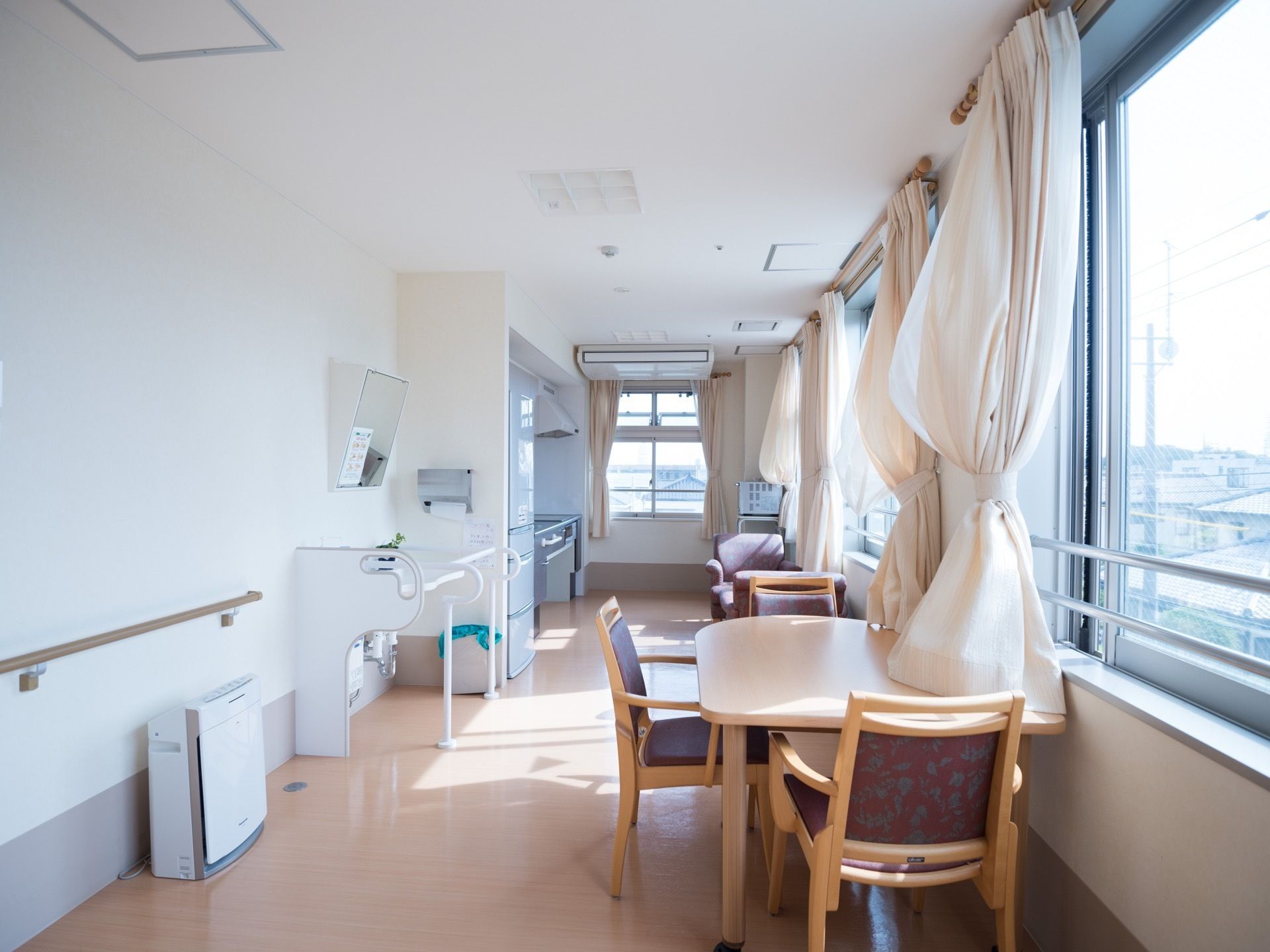 大きな窓に沿ってテーブルと椅子が置かれている。壁際には冷蔵庫と洗面台、キッチンがあり、壁には手すりが設置されている。