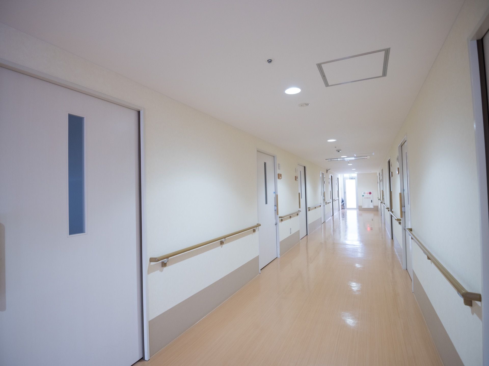 白い壁とドアの並んでいるベージュの床の廊下がある。ダウンライトの柔らかな光で照らされ、壁には手すりが設置されている。