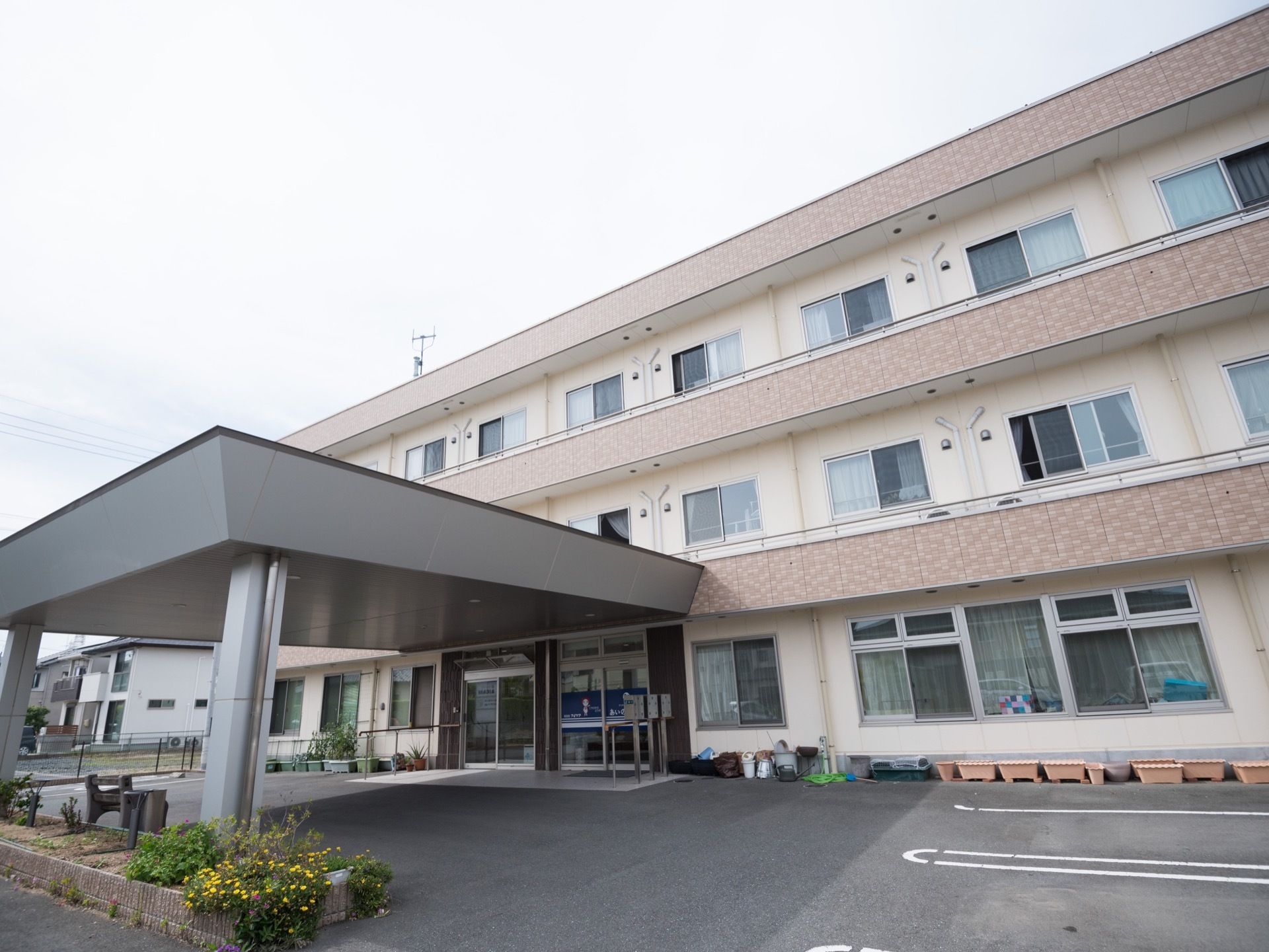 ベージュとクリーム色の壁の３階建ての大きな建物。玄関先は大きなルーフで覆われている。植え込みがあり、建物に沿ってプランターが置かれている。