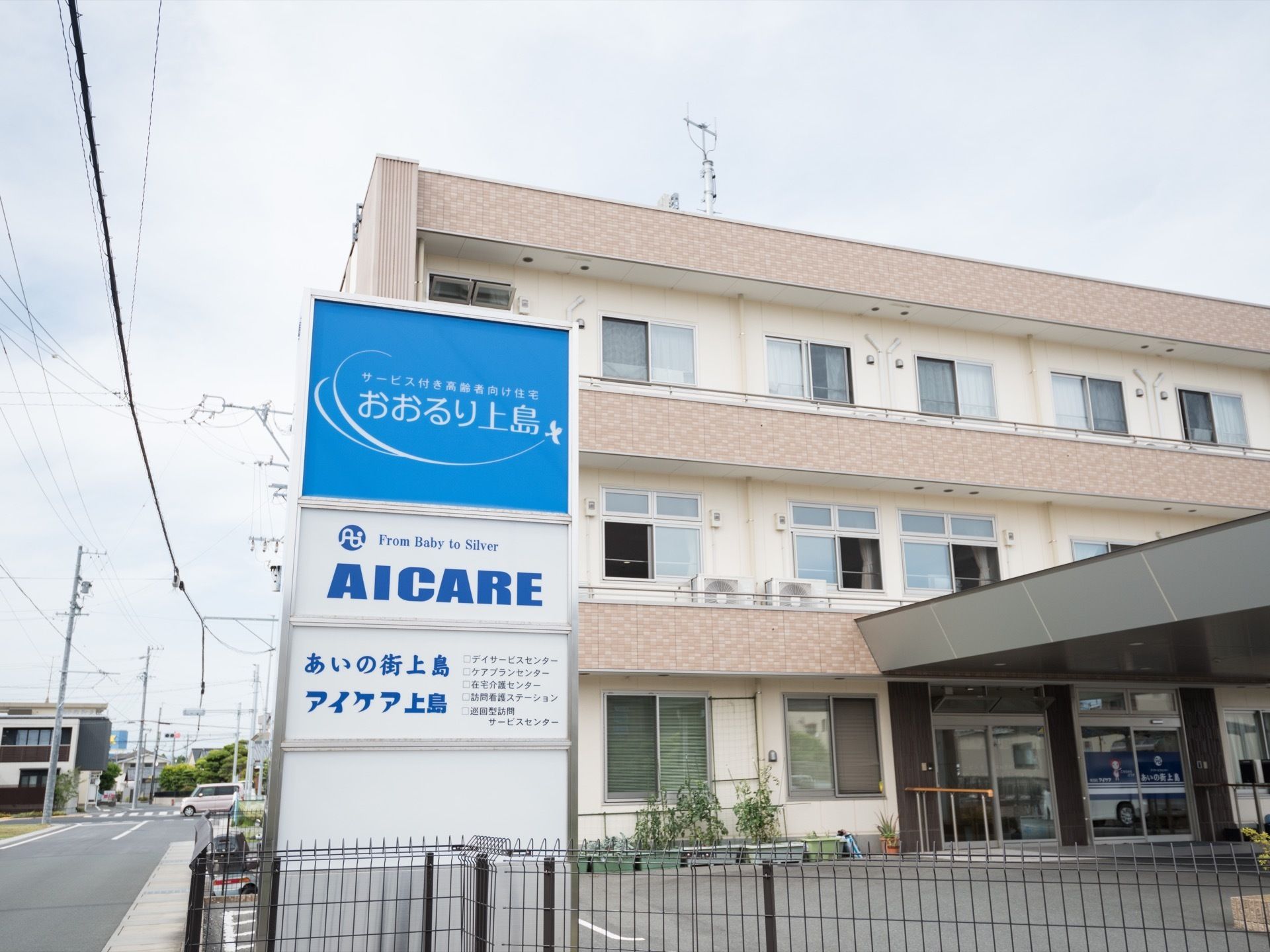 道路に面した場所に３階建ての大きな建物がある。周囲はフェンスで囲まれており、道路沿いに青い大きな看板が立っている。