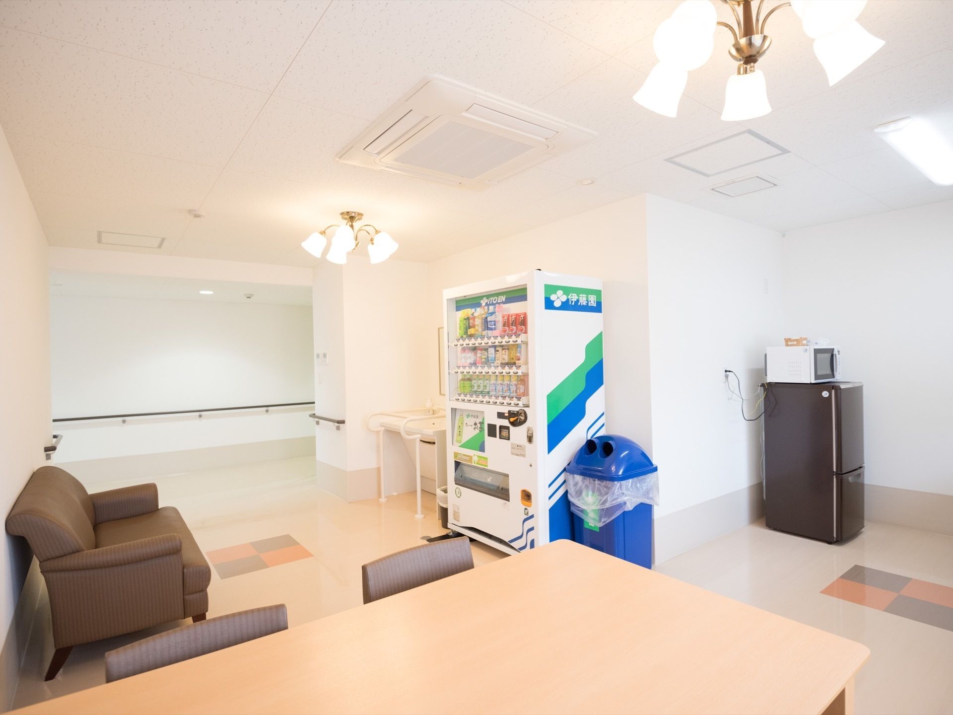 テーブルと椅子の近くにはソファーがある。ソファーの前には洗面台と自動販売機が設置されている。黒いミニ冷蔵庫と電子レンジも置かれている。