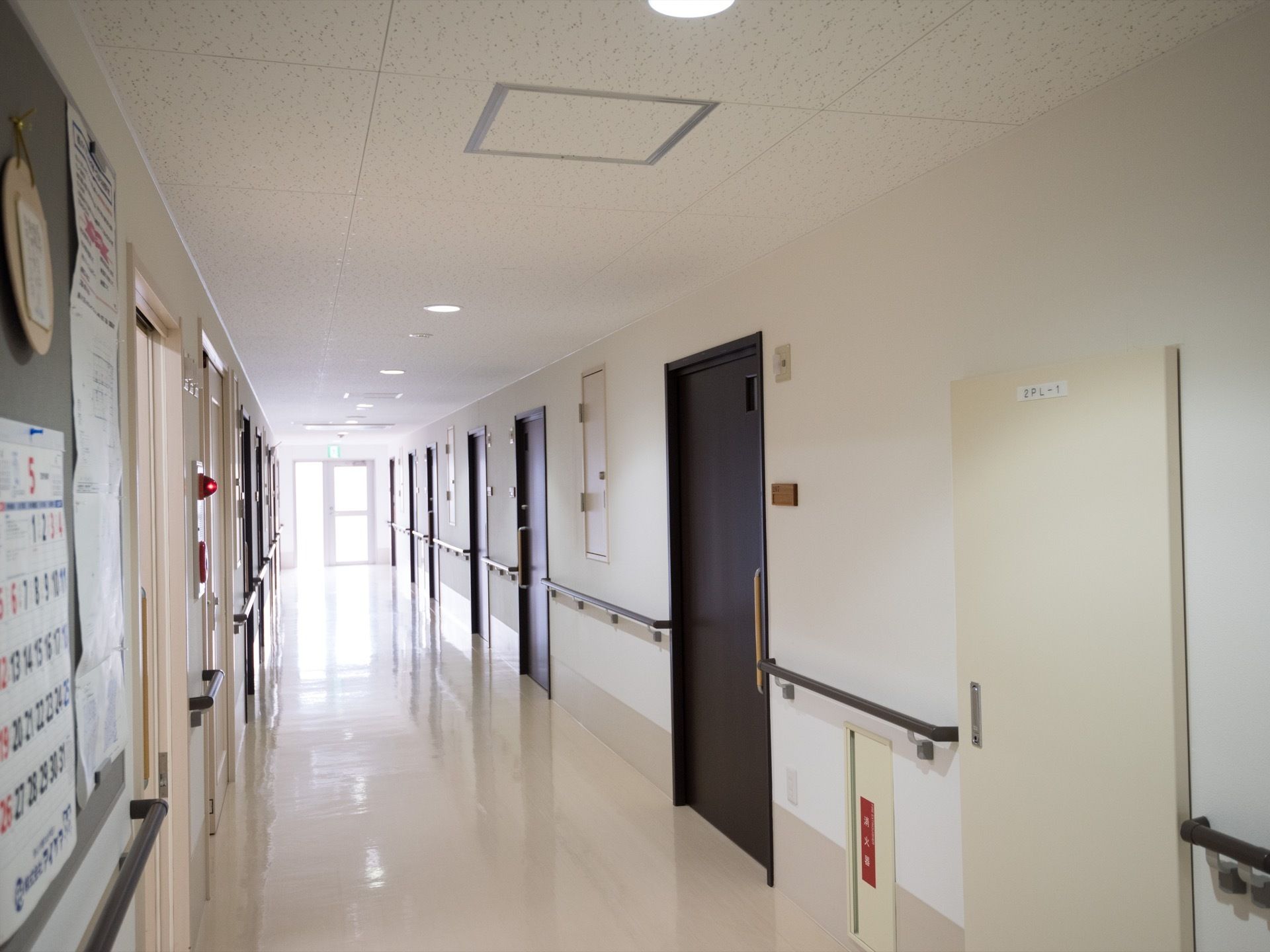 通路には部屋の扉があり、壁には手すりが取り付けられている。掲示板もあり、カレンダーや紙が貼られている。