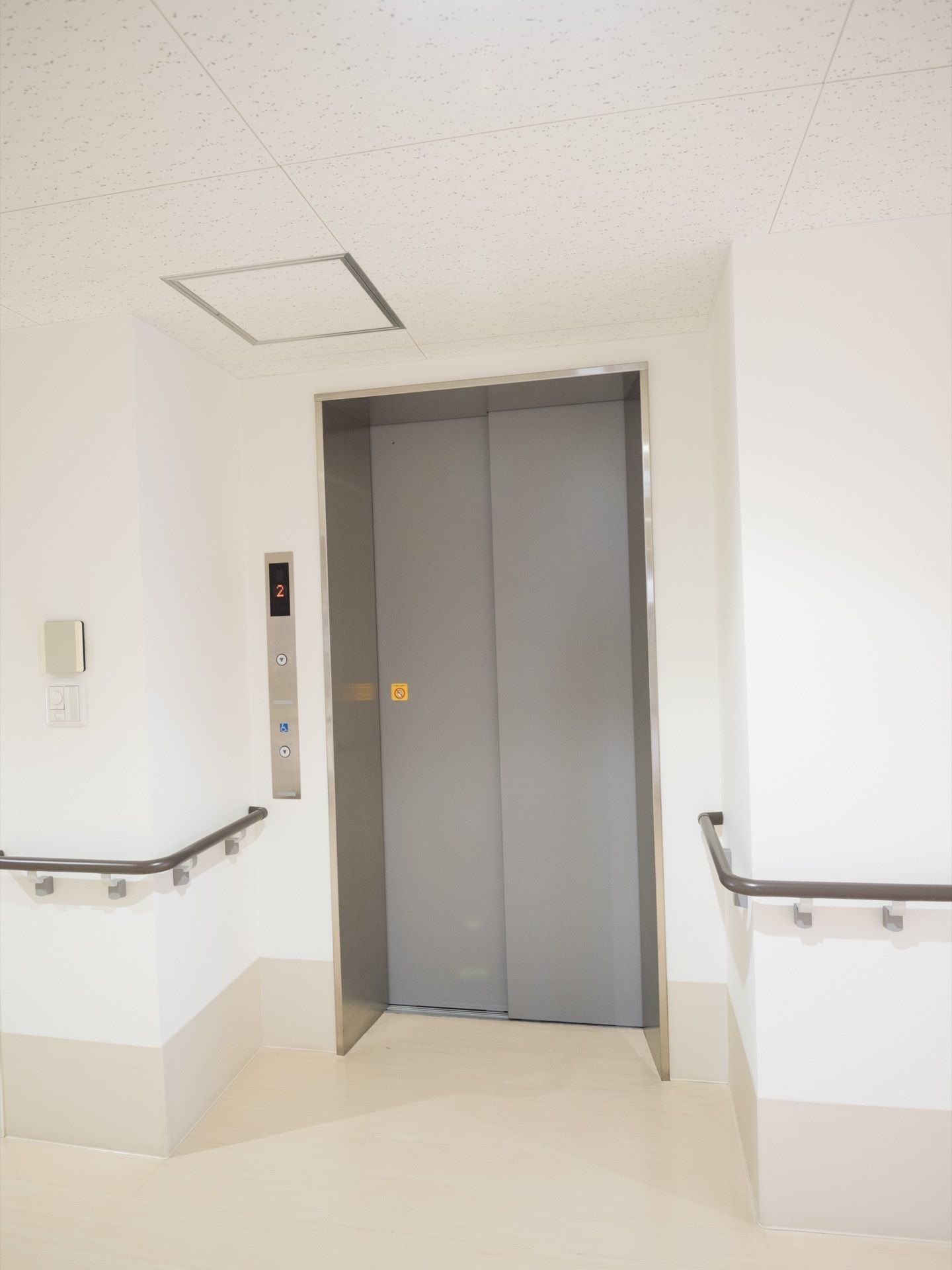 白色の壁、薄いベージュ色の床がある。そこにグレー色の扉のエレベーターが1基設置されている。壁には、手すりが取り付けられている。
