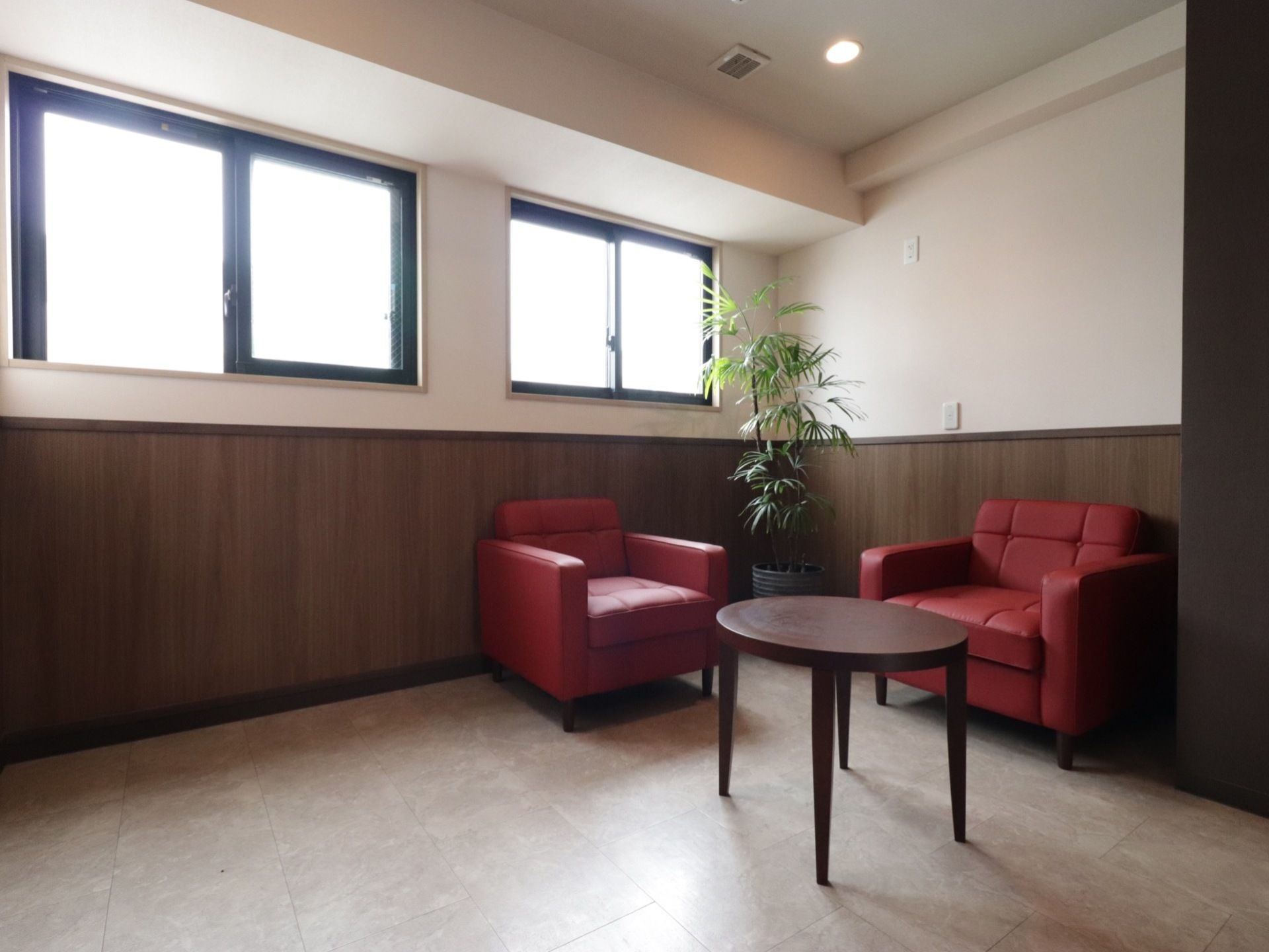 茶色の丸いテーブルと一人掛けの赤いソファーが２脚置いてある。２つの窓が設置され、観葉植物が飾られている。