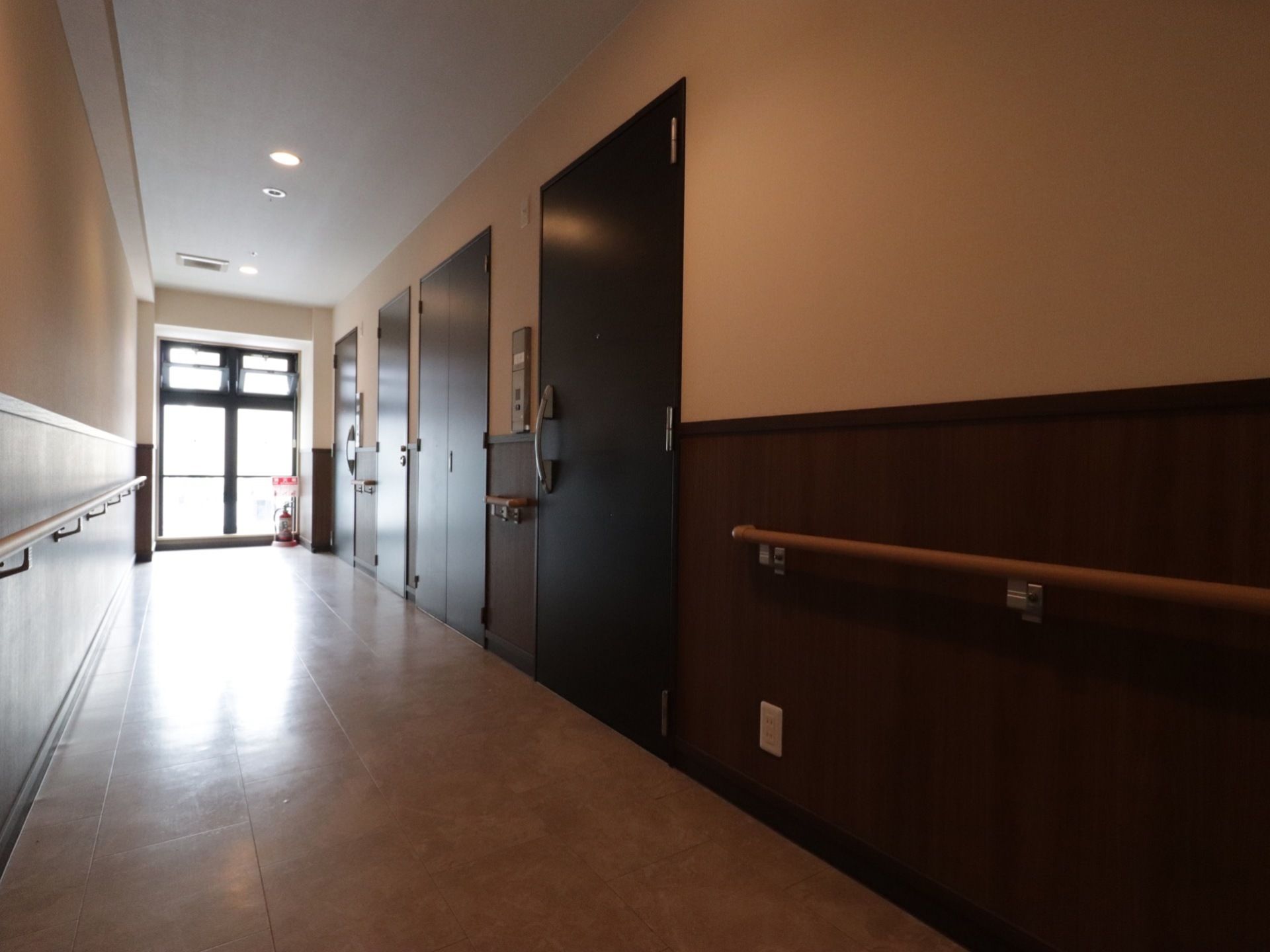 通路の両側に手すりが設置されている。通路の先に大きな窓があり、光が差し込んでいる。窓際に消火器が置いてある。